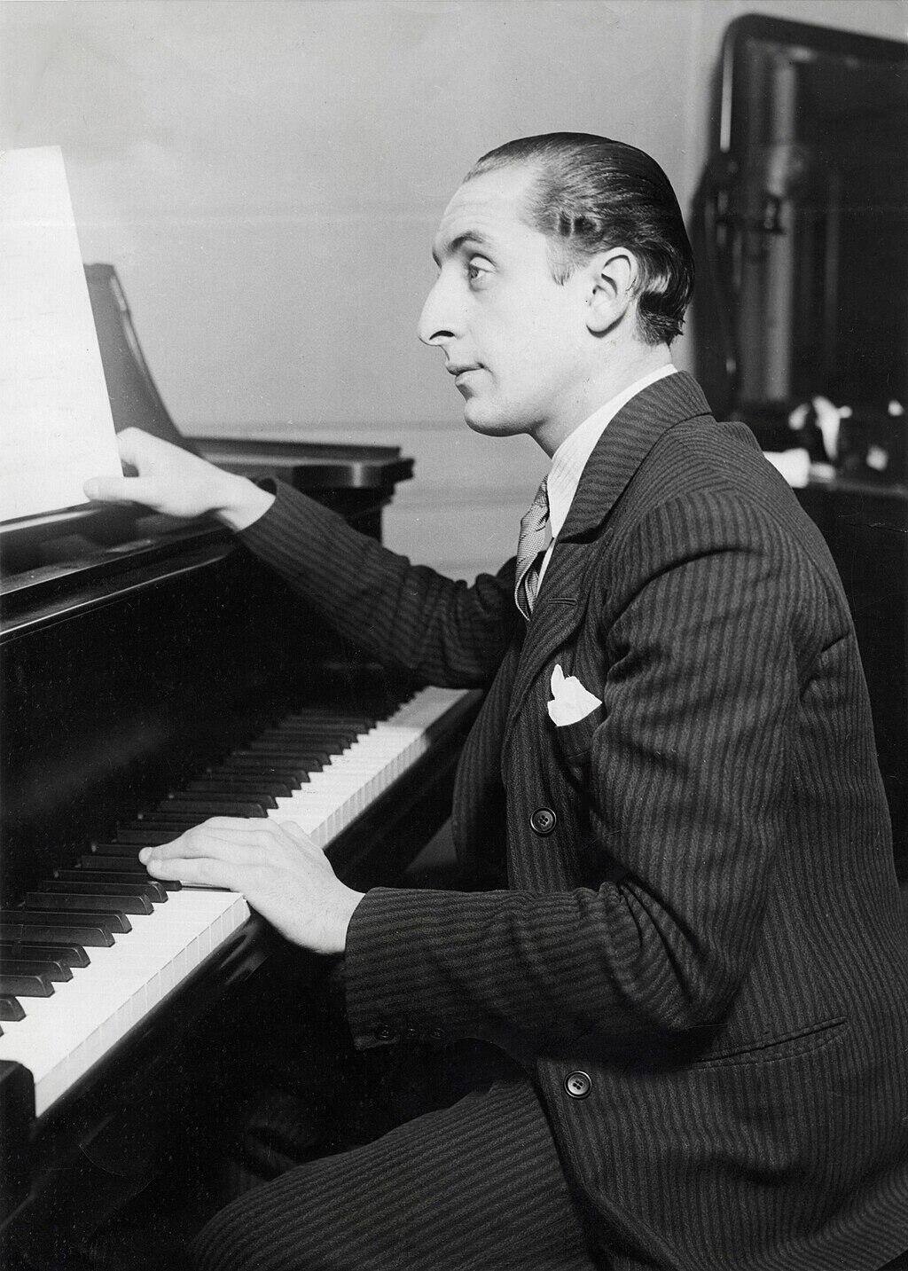 Vladimir Horowitz sits at a piano, his right hand poised on the keys while he reads from sheet music on the stand.