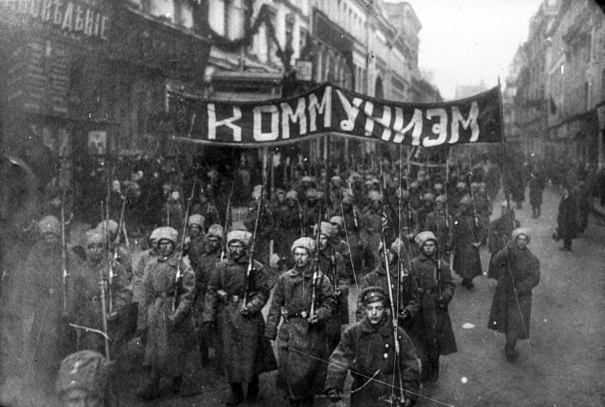black and white photo of a mob of soldiers marching in the streets of Russia holding a big sign reading "Communism" written in Russian