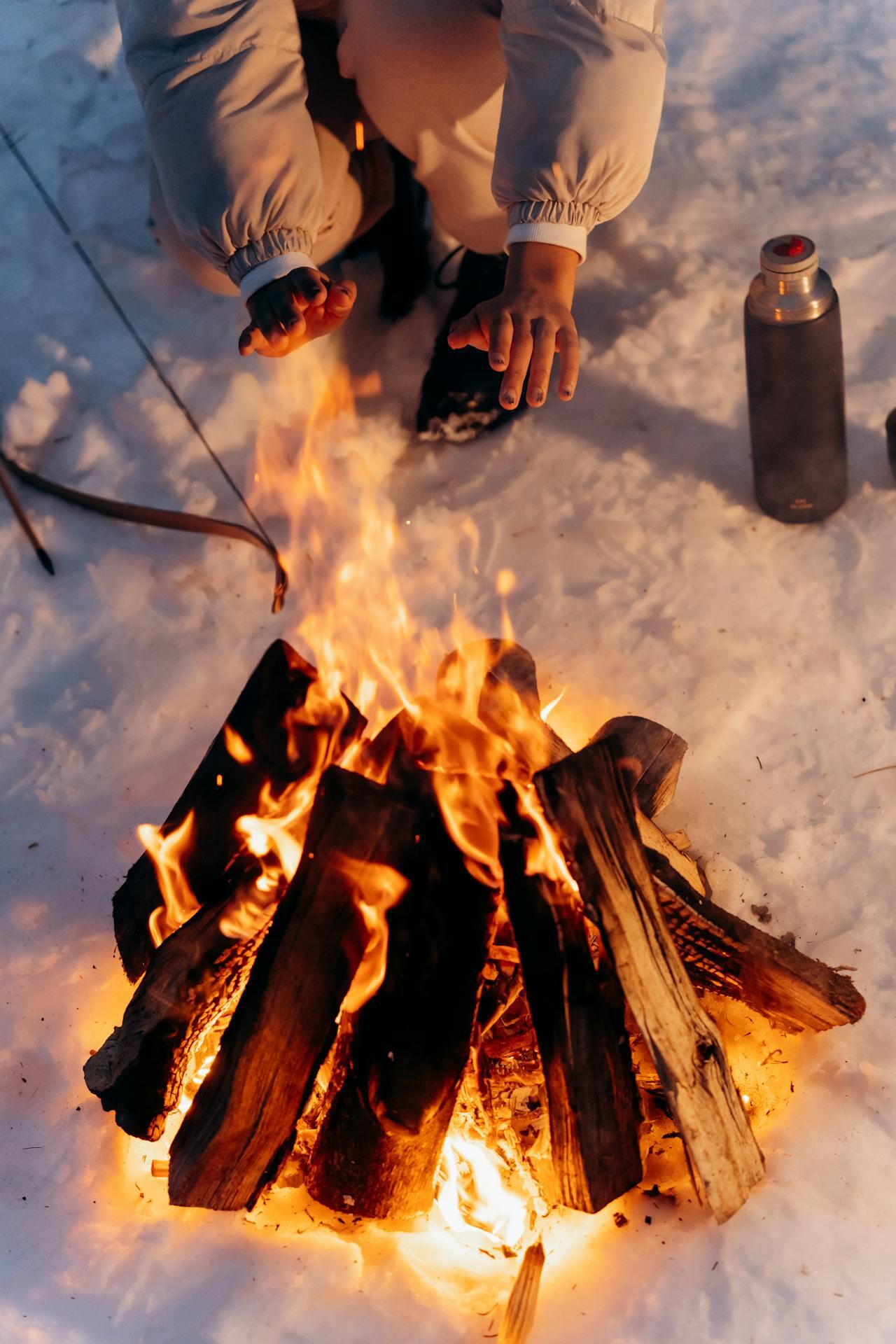 person in the snow warming their hands with a campfire. A bow and arrow and bottle of water are in the background