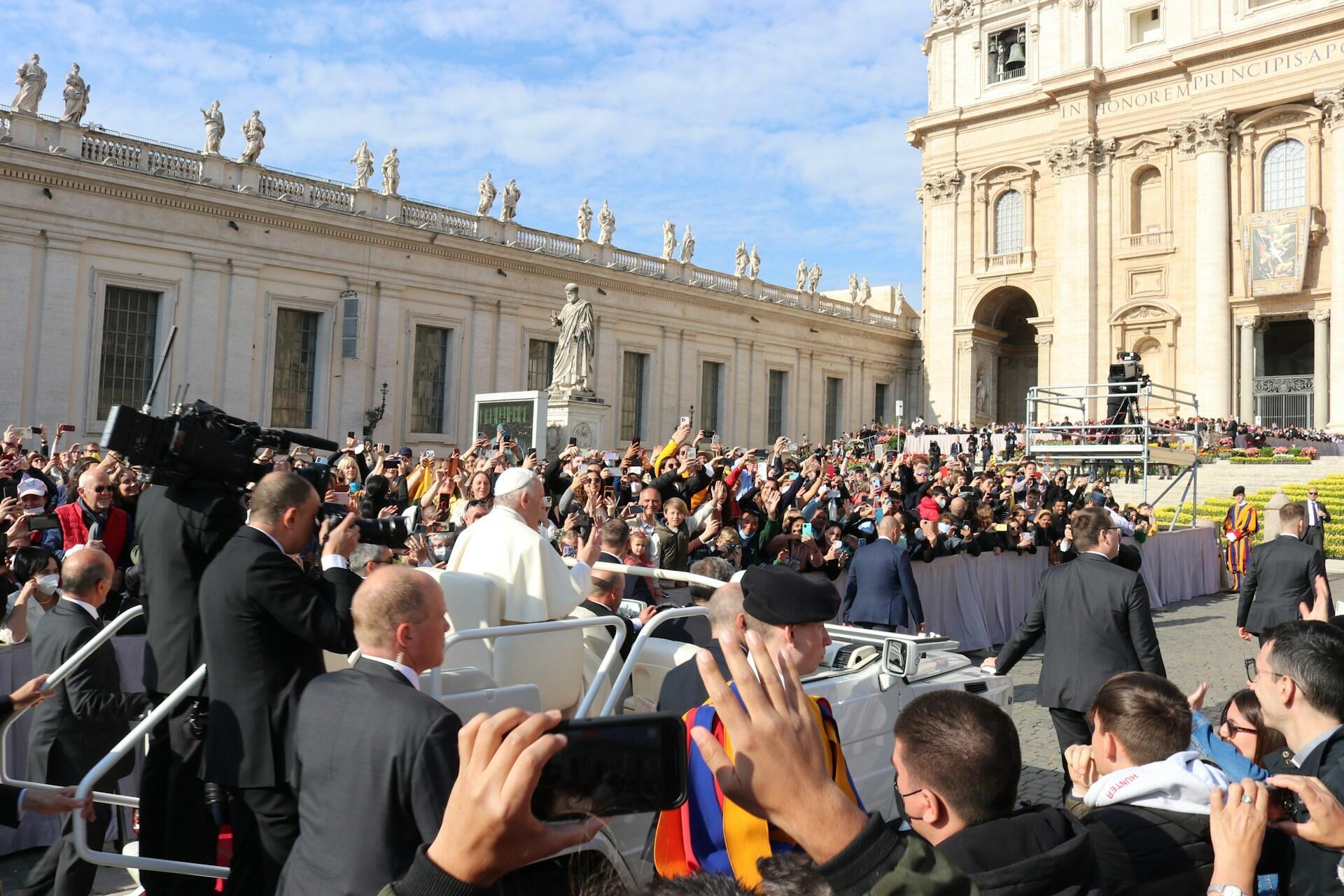 A crowd gathers excitedly around the Pope, capturing the moment with smartphones near St. Peter's Basilica on a sunny day.