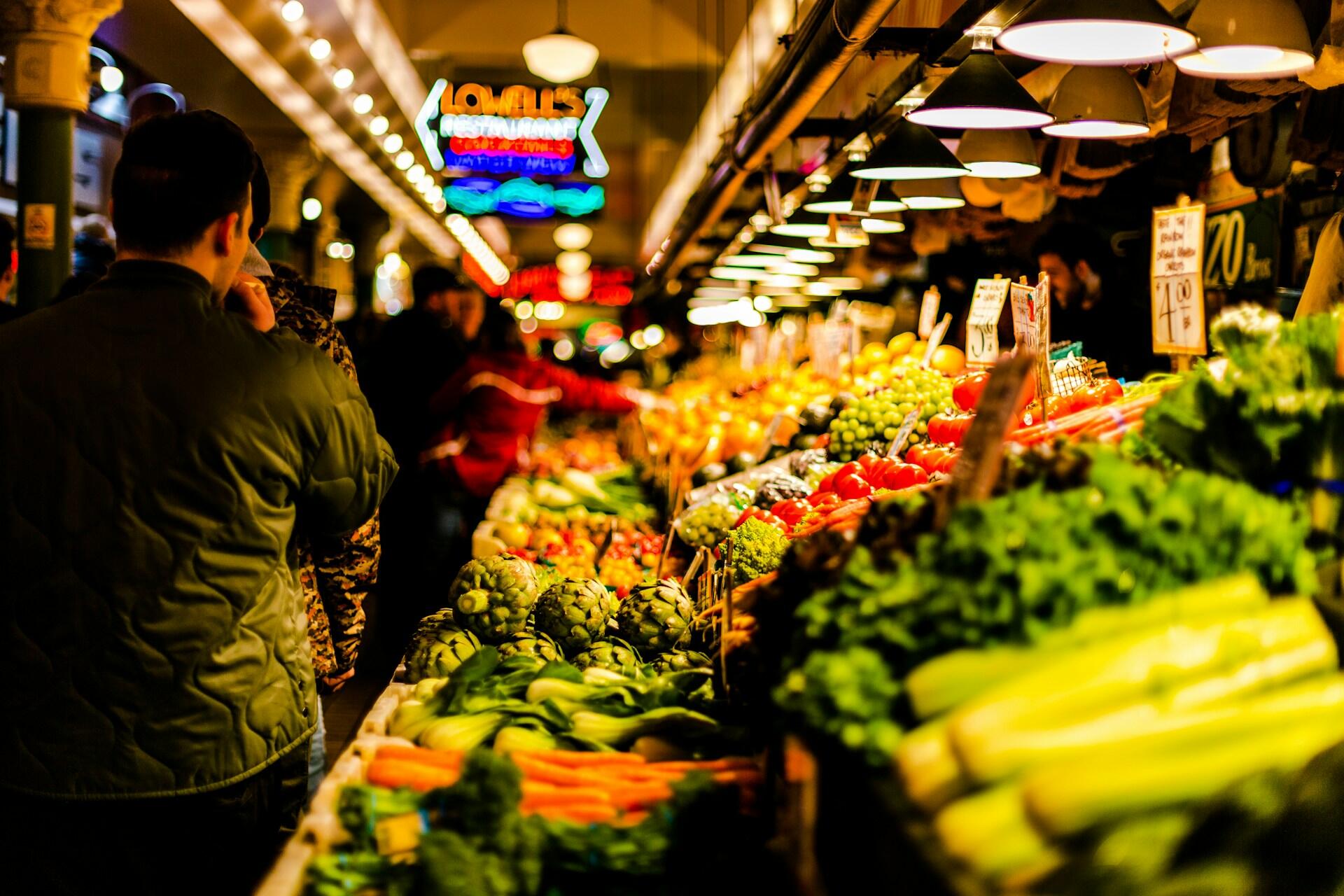 People browsing fresh fruit and vegetables at a brightly lit indoor market stall.