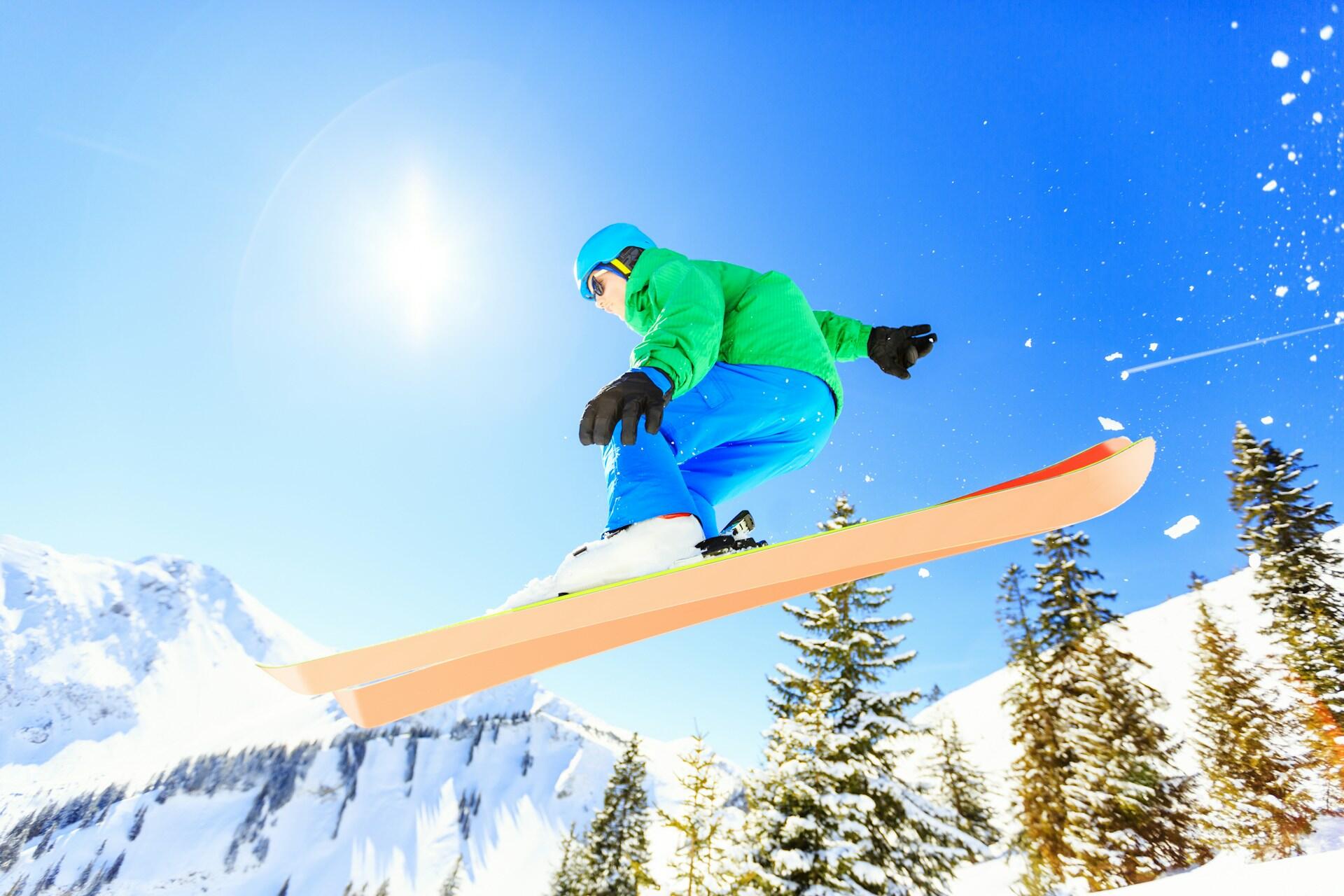 A man skiing in the air on orange skis wearing a green jacket and blue snowpants.