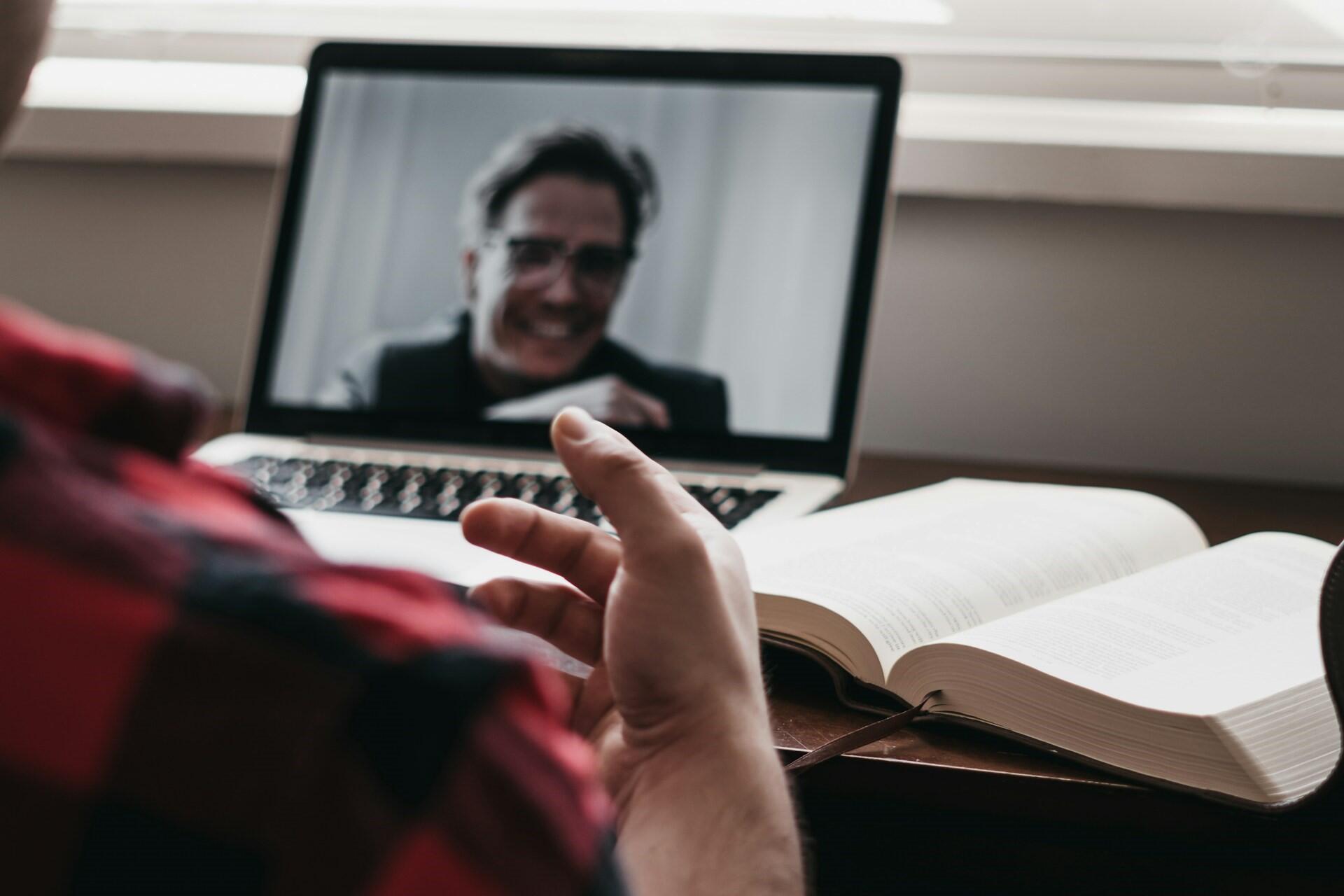Student attending an online lesson on a laptop while reading from an open textbook.