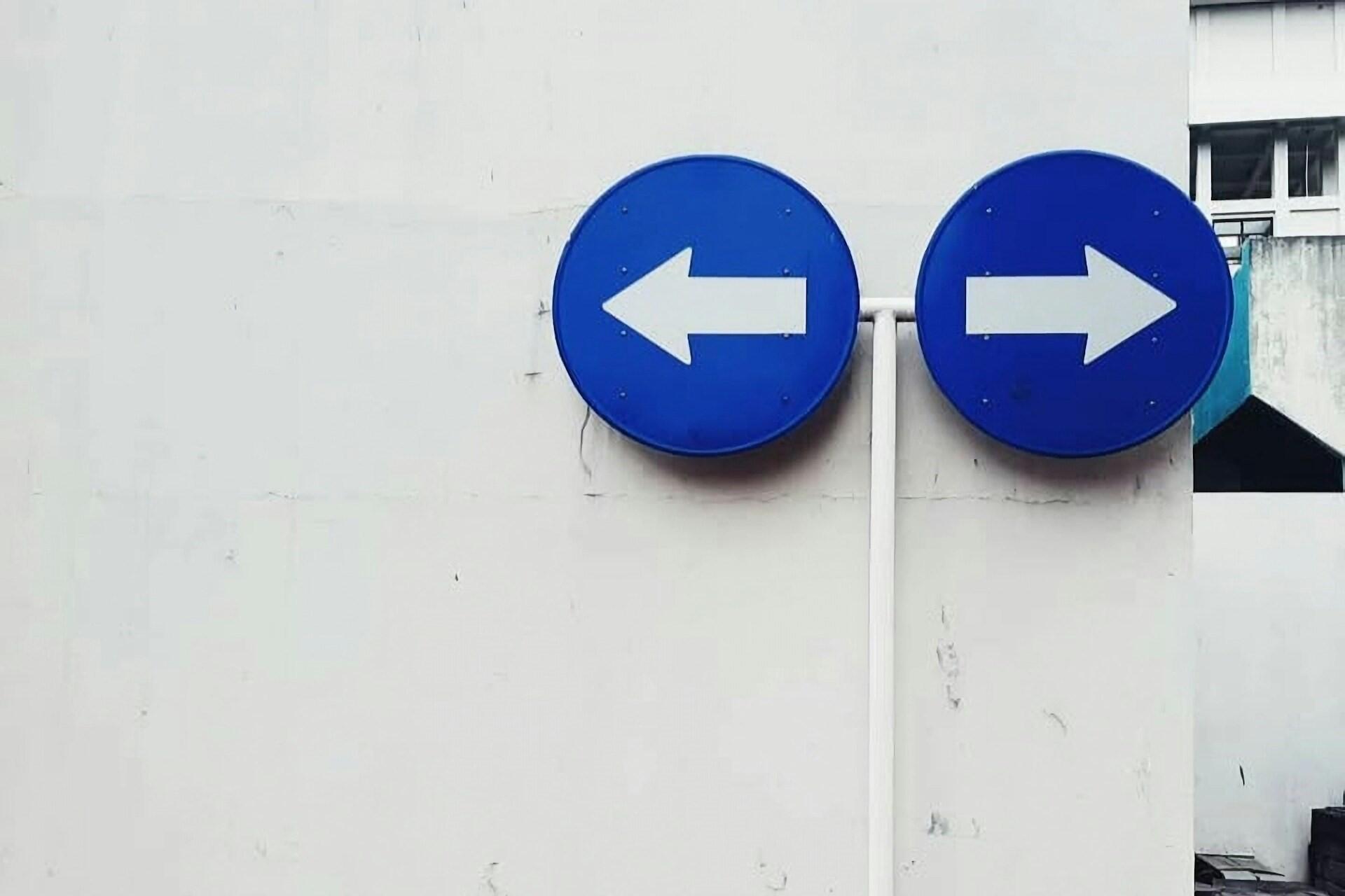 Two blue circular road signs with white arrows pointing left and right mounted on a white wall.