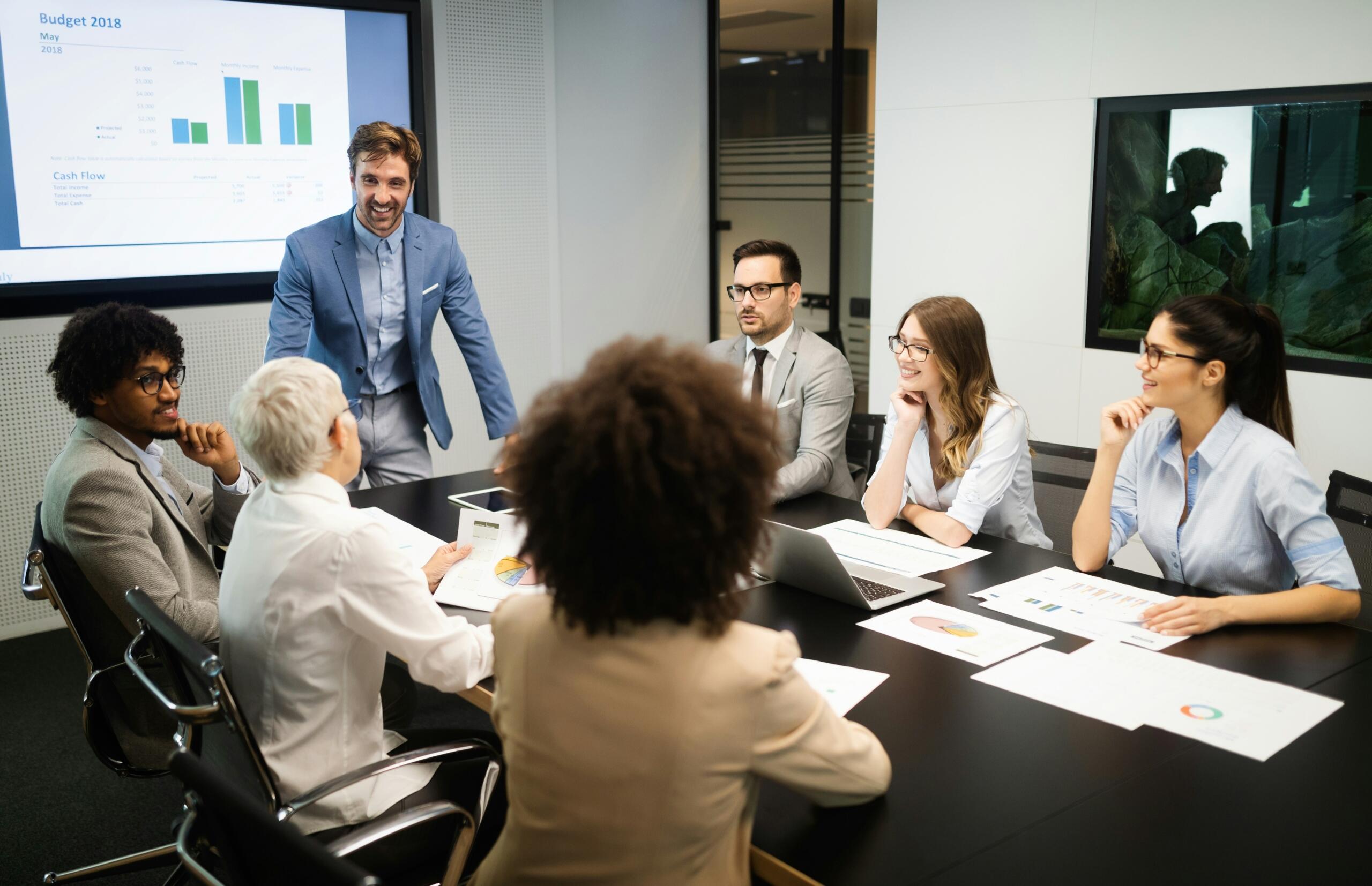 Seven people in a business meeting with one presenter discussing the 2018 budget.