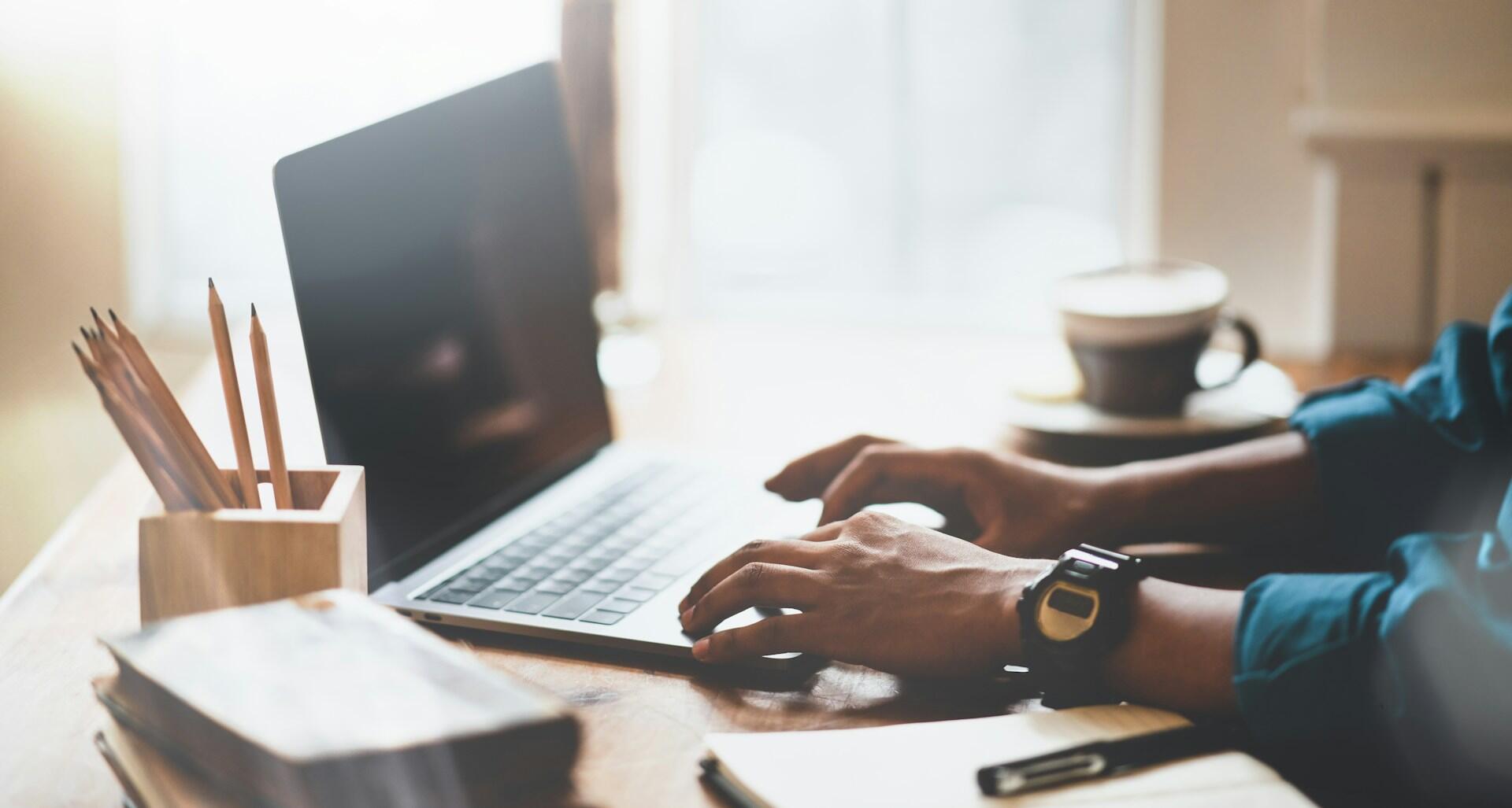 student typing on a laptop computer with pencils and notebooks in the foreground and a cup of coffee in the background