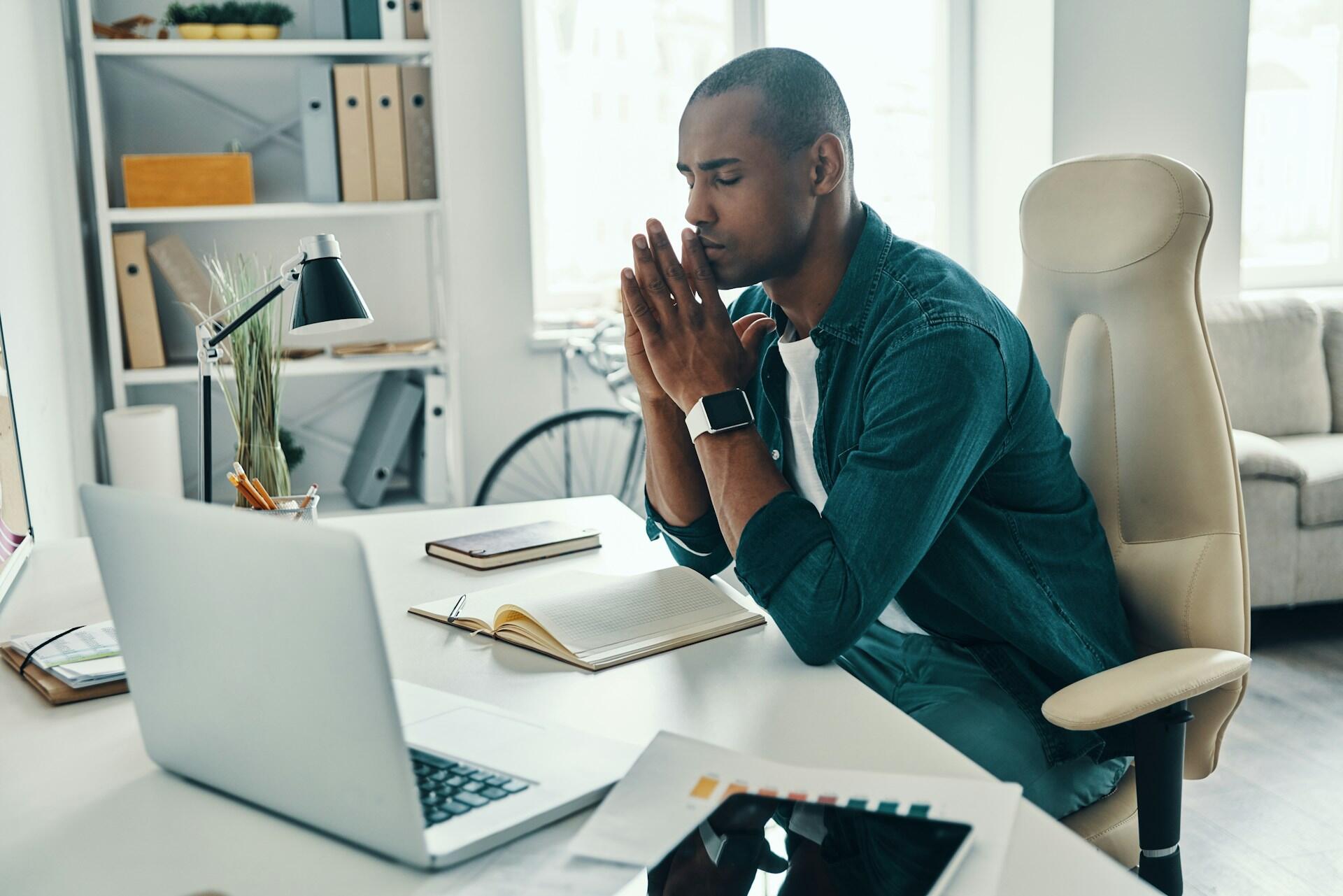 man sitting at a desk with elbows resting on the top, palms together, eyes closed, in order to relax and focus