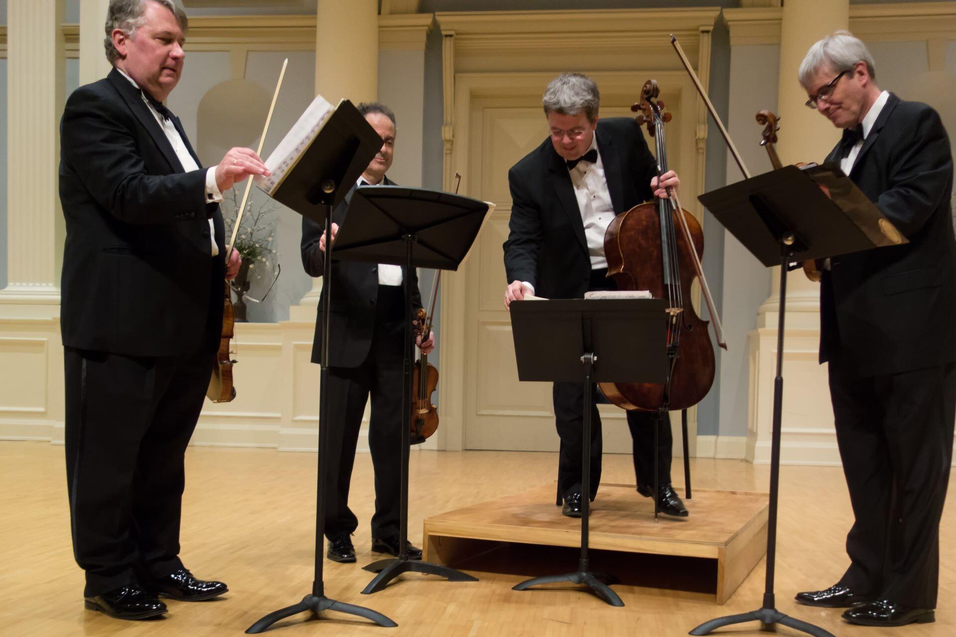four musicians in formal black suits standing on a concert stage with violins and a cello adjusting sheet music on stands