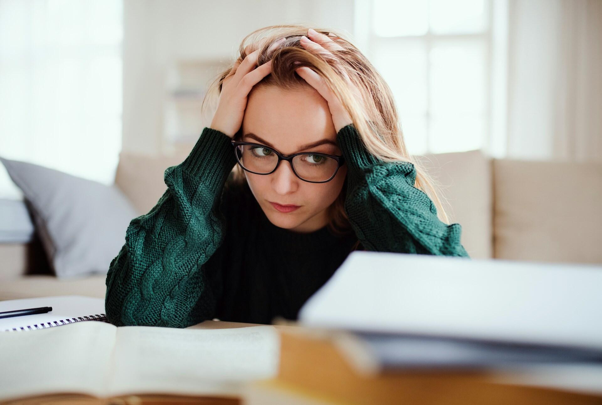 woman appearing overwhelmed, her hands in her hair, while looking at piles of books and notes