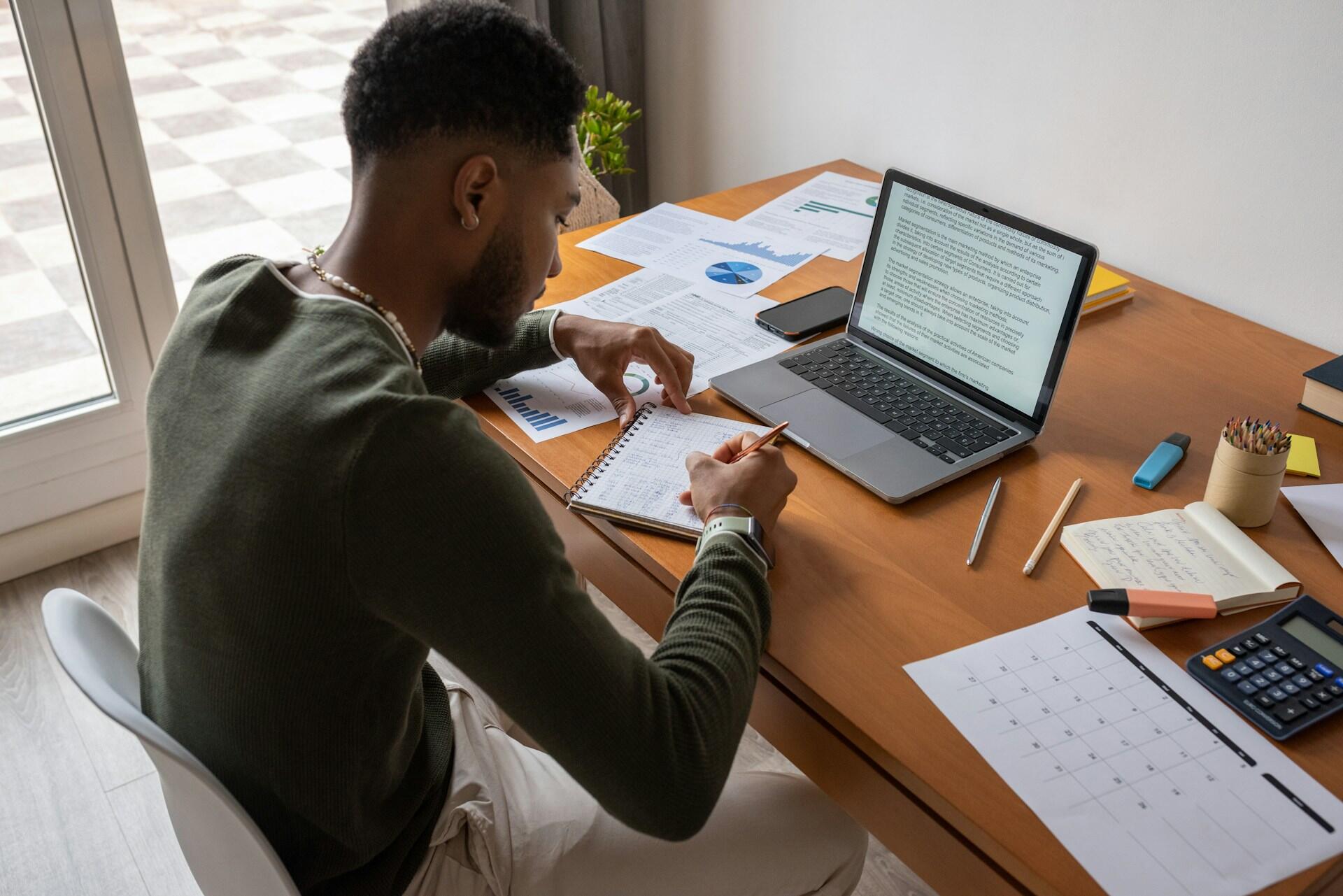 person writing notes while looking at text on a laptop screen. A calendar planner and printed charts are nearby