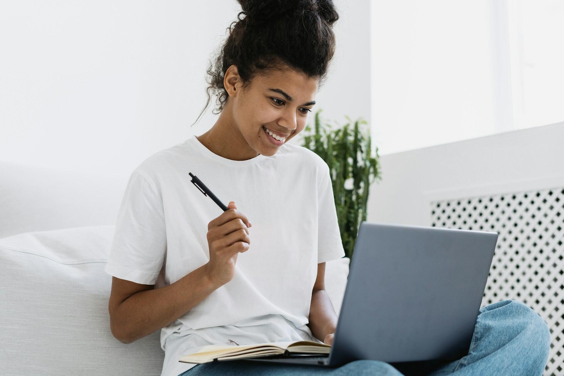 Student looking at a computer screen, smiling, and prepared to take notes with a pen and notebook