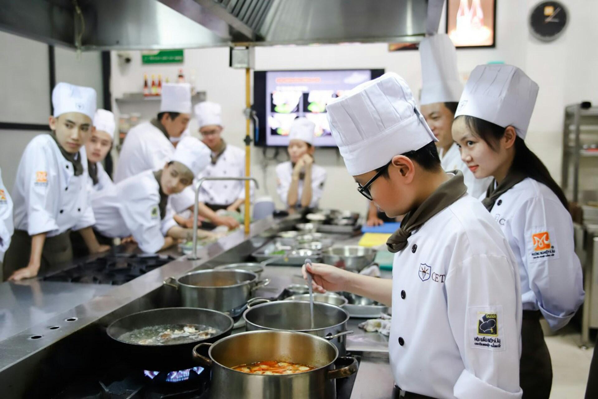 Nine chefs in white coats and hats cooking in a cooking class.