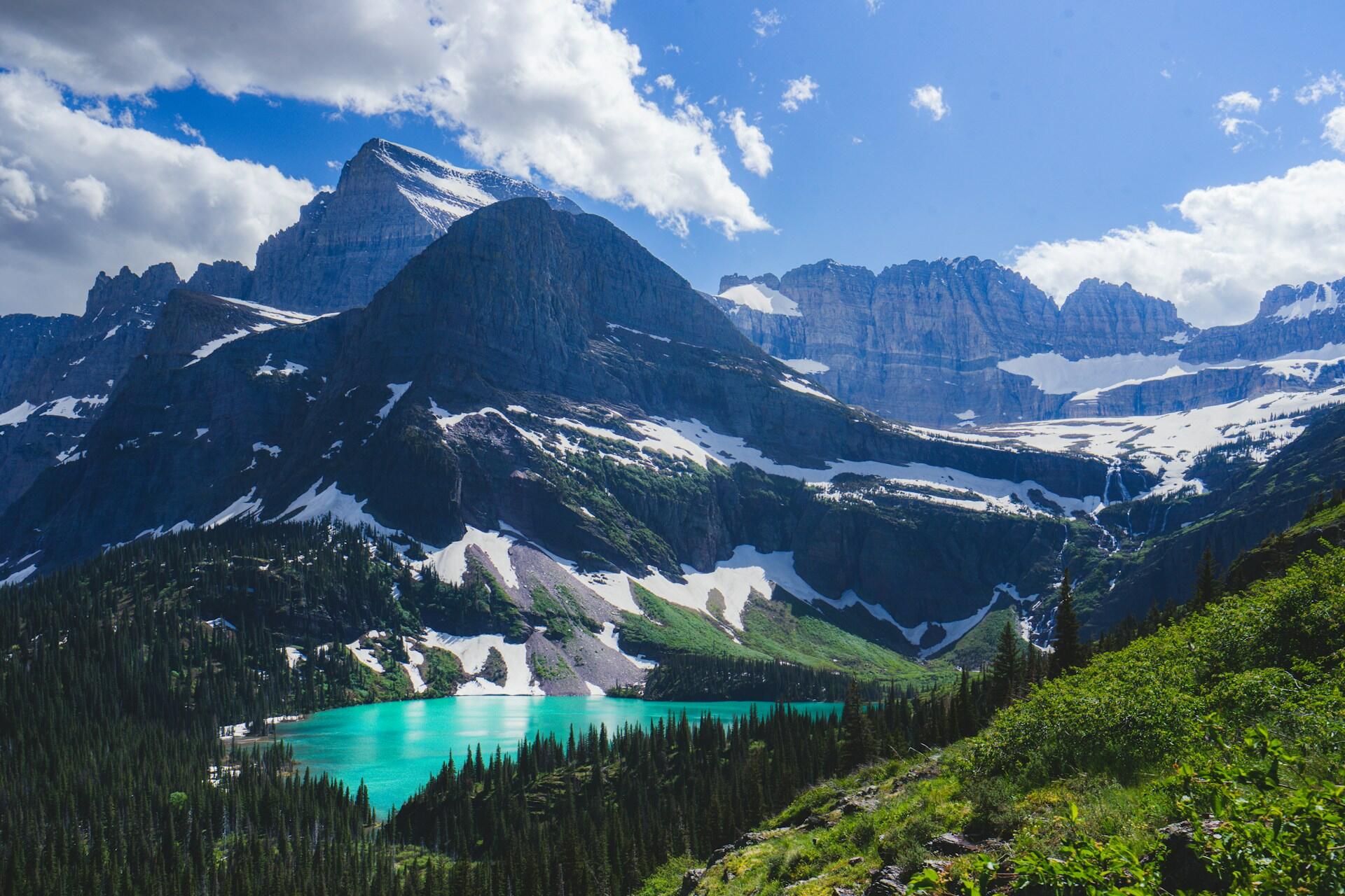 a landscape with green hills and an aquamarine lake in the foreground, a high, steep hill in the middle ground, and rocky, jagged peaks in the background