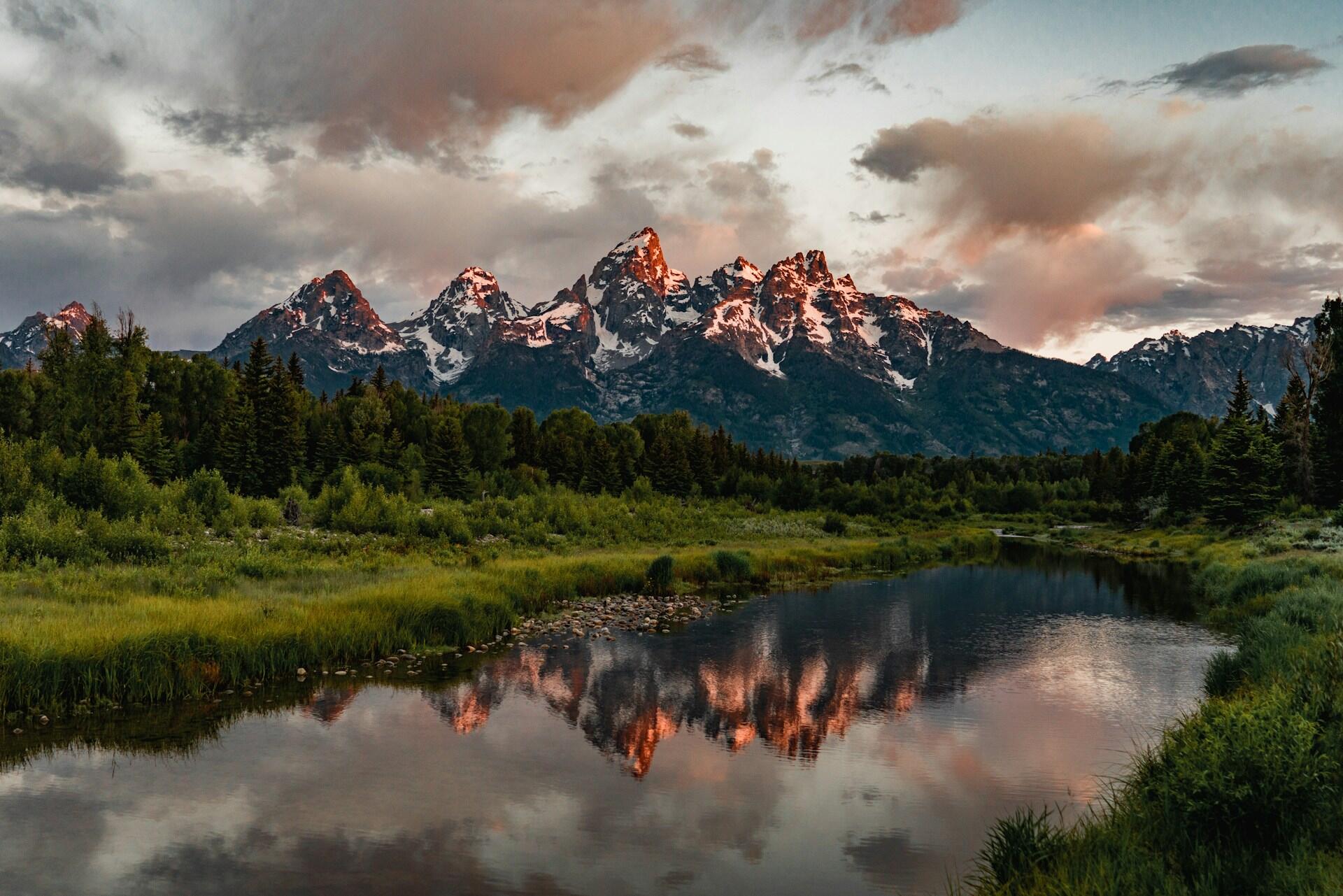 view of faraway snowcapped sharp mountain peaks with a thick forest and clear lake in the foreground
