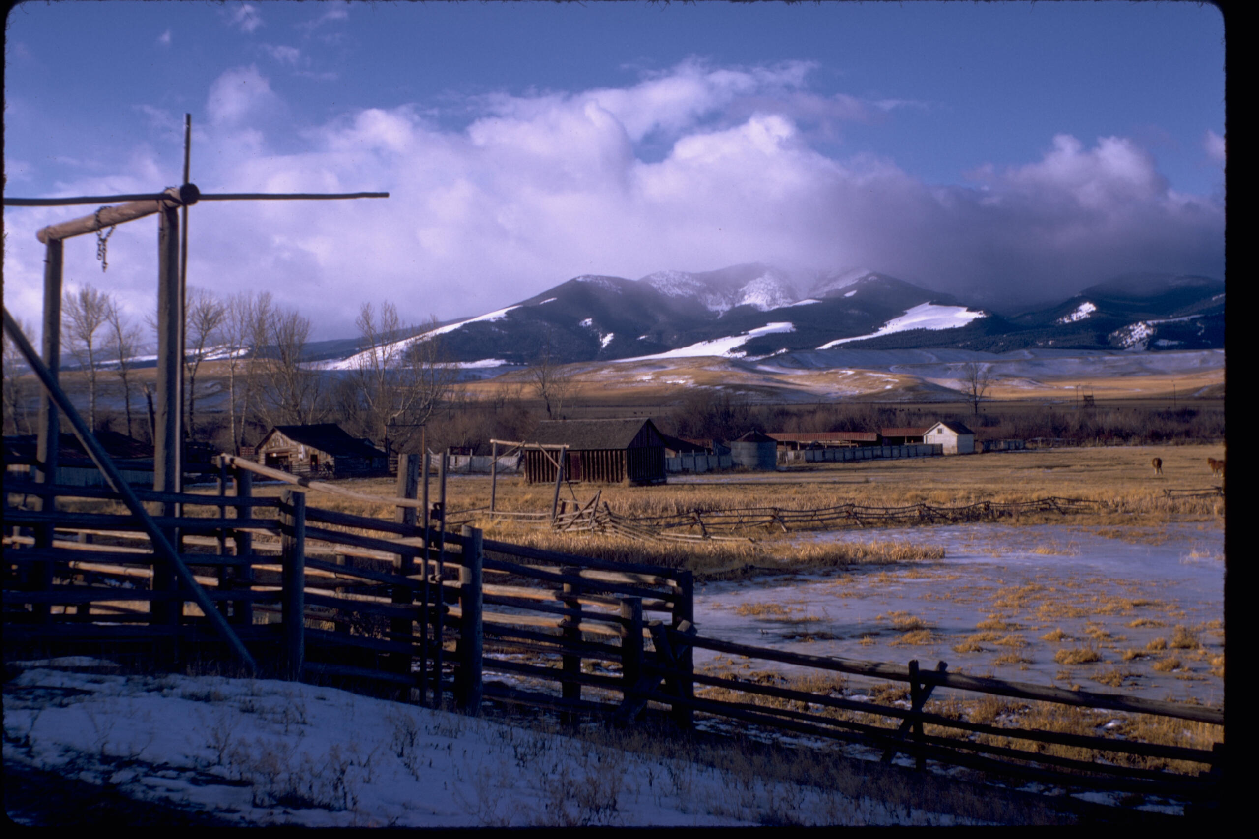 a picturesque view of the ranch, with small amounts of snow and an icy pond with flat prairie area surrounding, with gentle hills backed by tall, snowy mountains and low clouds in the background
