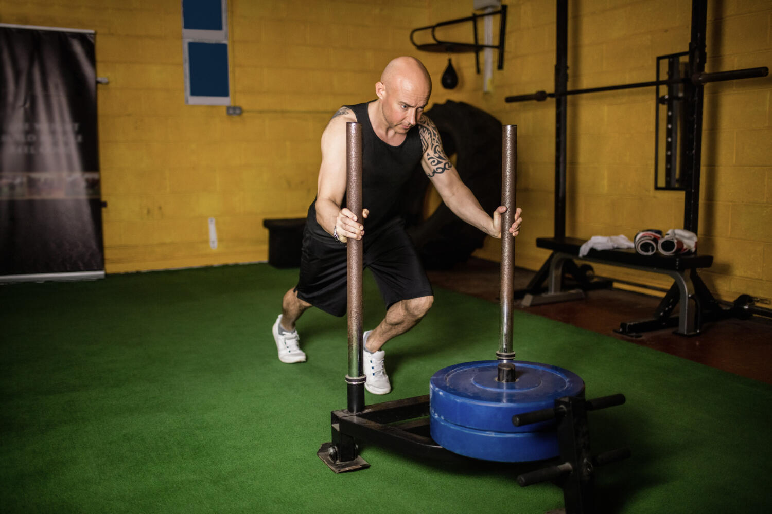 A person performs a sled push in a gym, showcasing strength training on a green gym floor with yellow walls in the background.