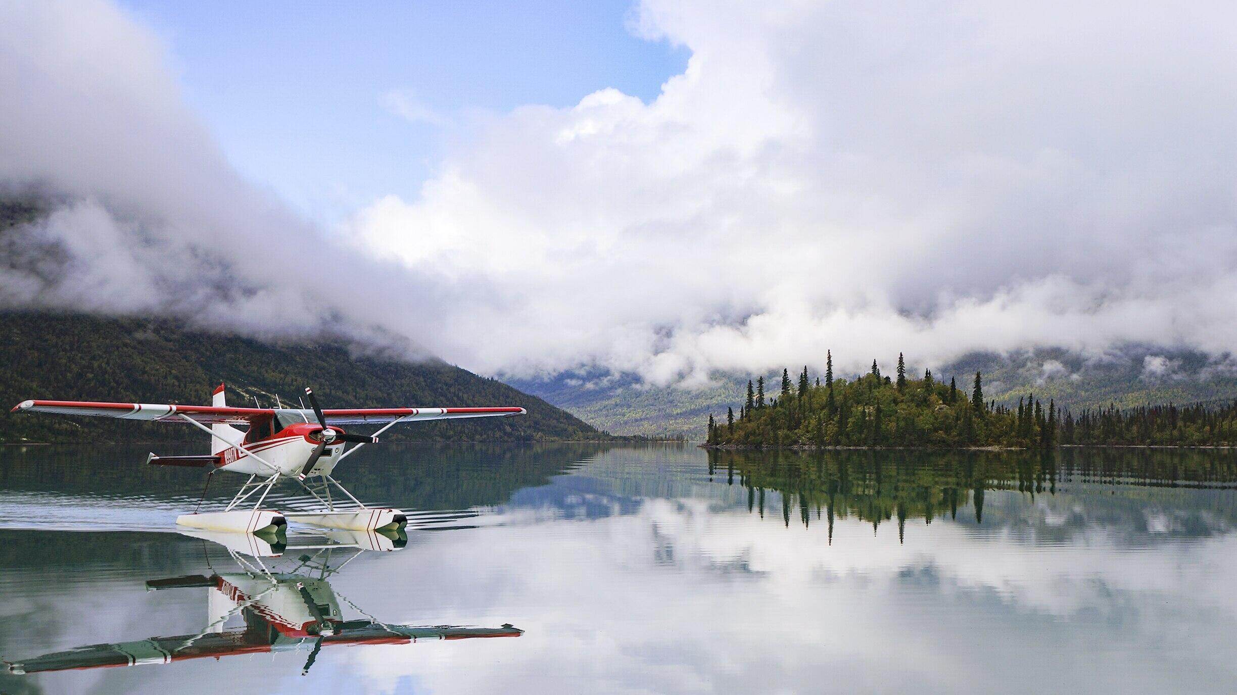 enormous lake with still water reflects the sky full of low clouds, obscuring the nearby mountains. A red and white float plan coasts across the water