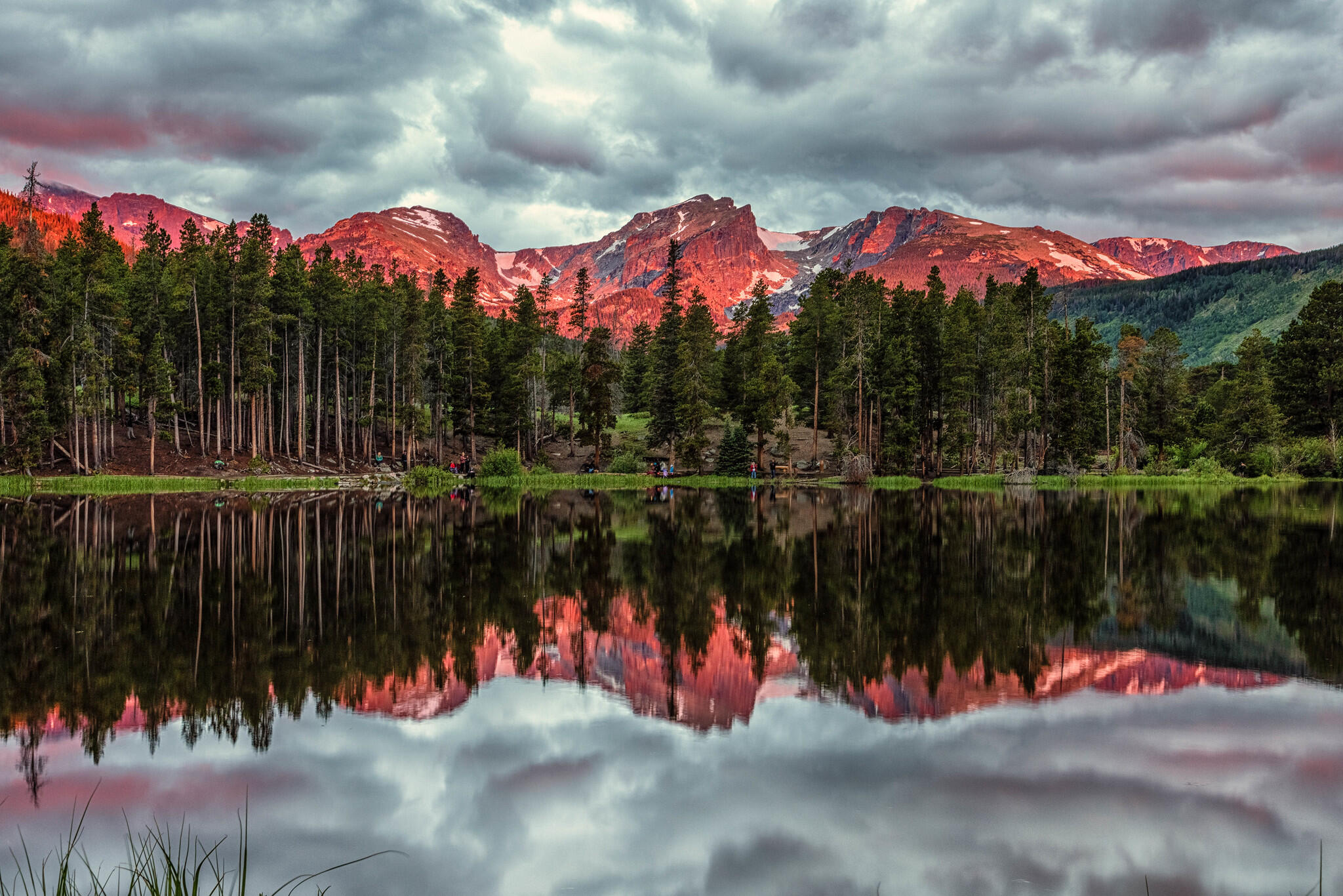 a still, clear lake in the foreground reflecting the Rocky Mountains, appearing a pinkish-red color in the sunrise light