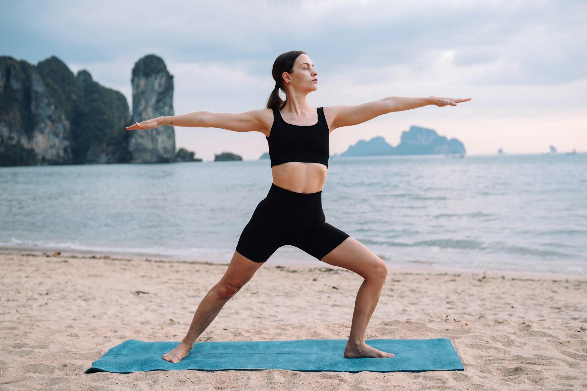 A girl in a black workout outfit on a blue yoga mat on the beach performing the warrior pose.