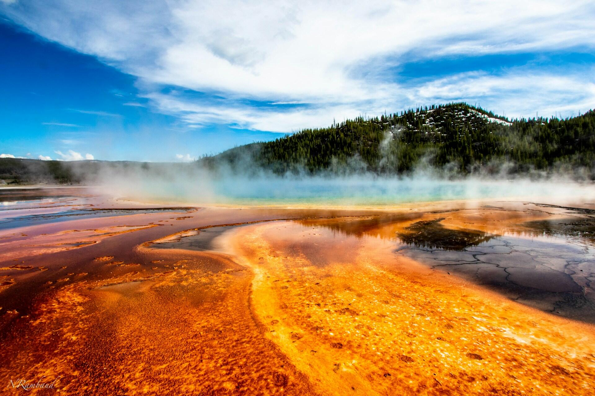 geothermal pools filled with bright orange, red-orange, and aquamarine algae and minerals with steam coming up off the water, slightly obscuring the forested mountain in the background