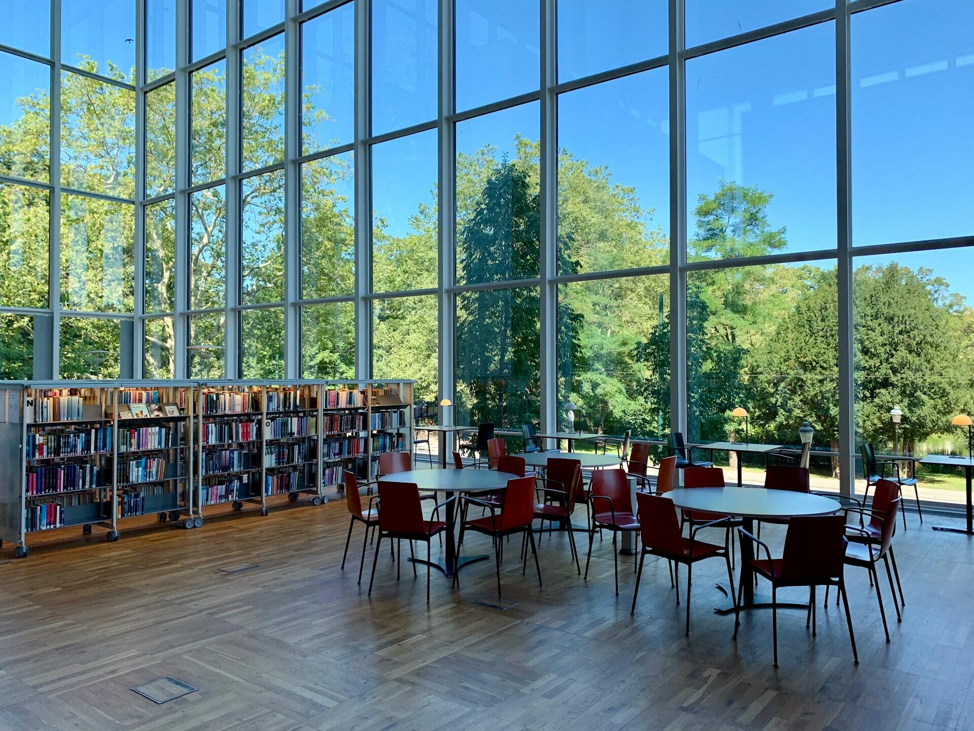 A school library with many columns of books, several round tables and floor to ceiling windows.