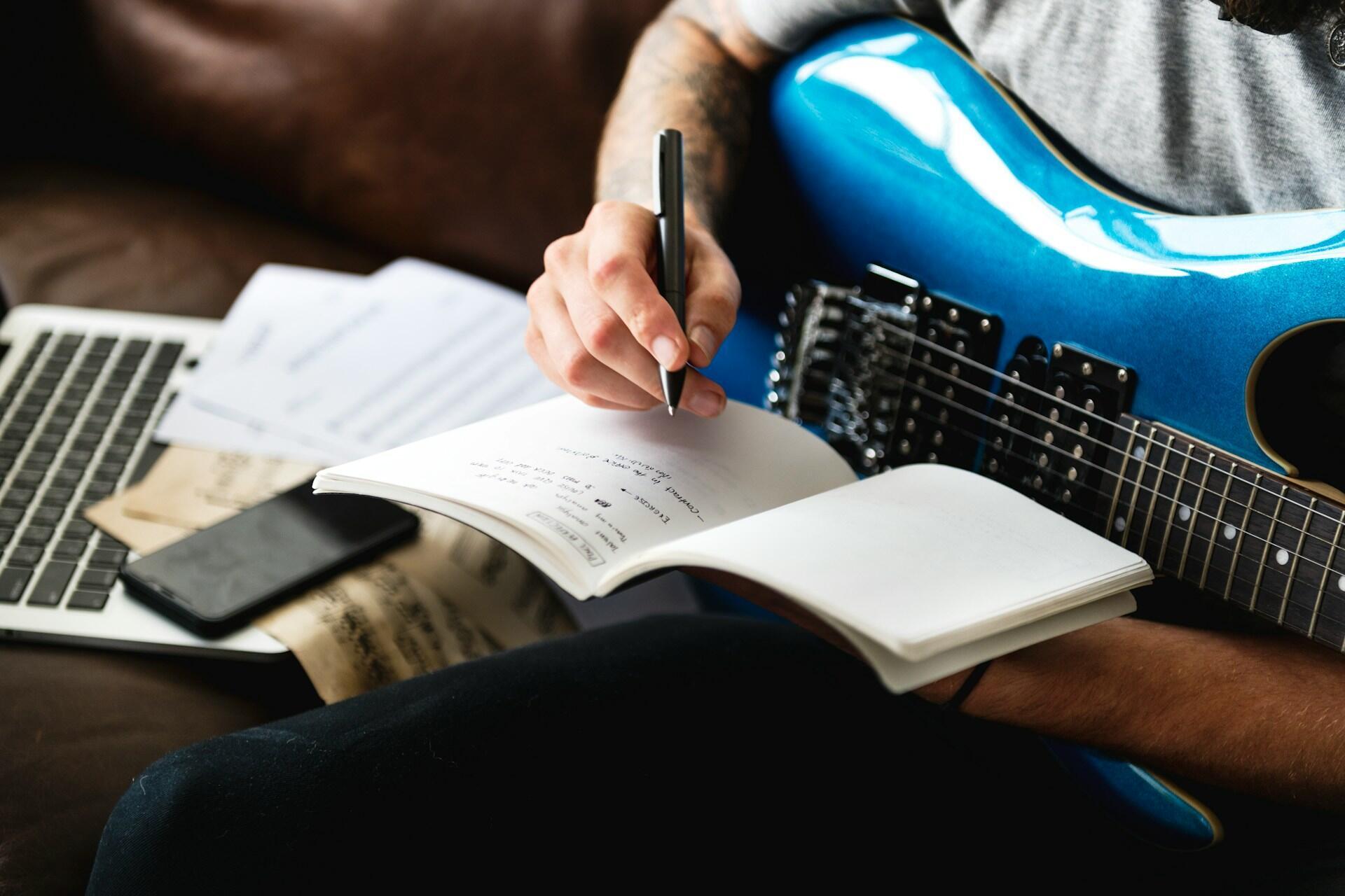 A guitarist writing down a song while playing a blue guitar.