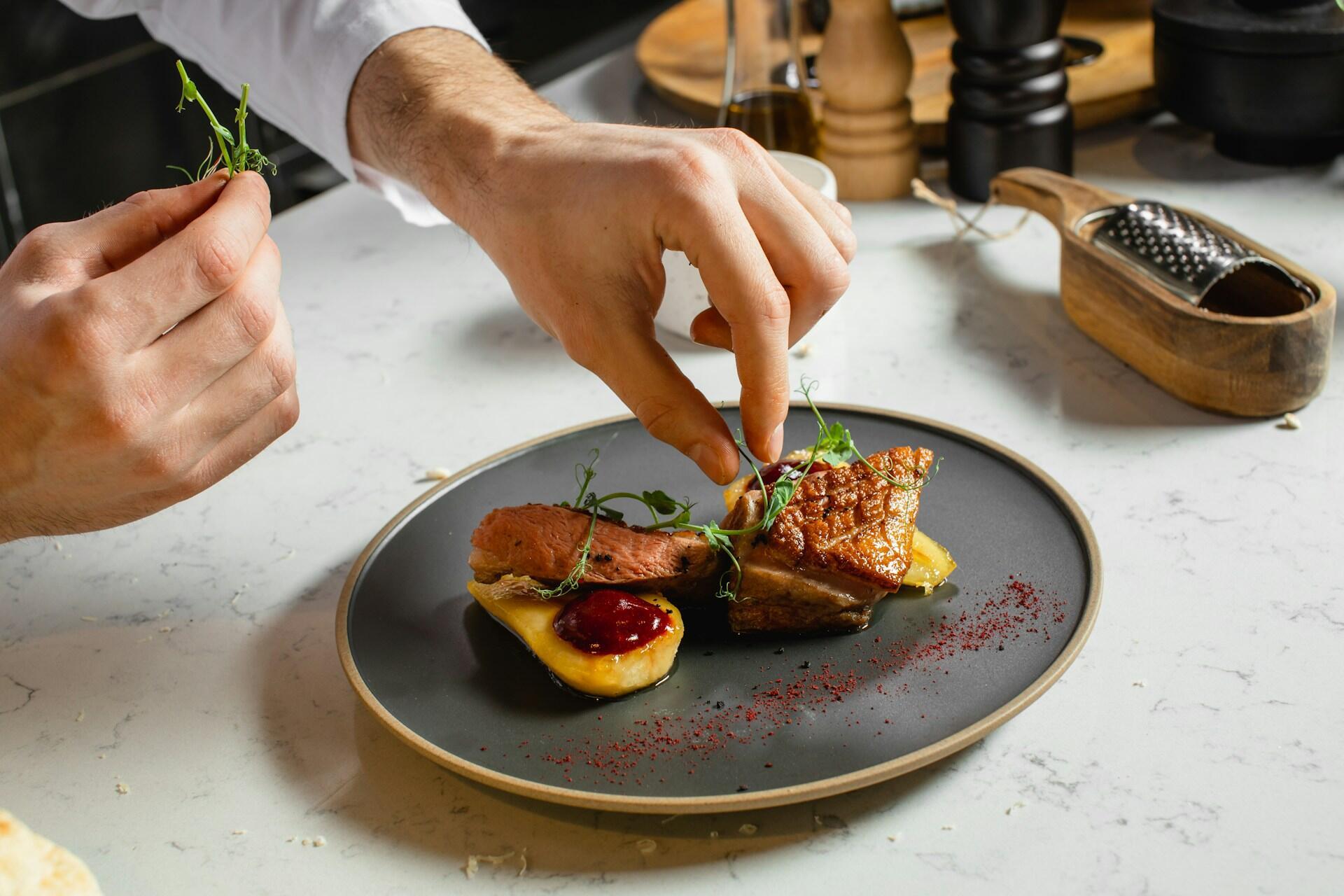 La mano de un chef decora un plato bellamente presentado con hierbas, con carne tierna y una salsa vibrante en un plato redondo y oscuro.