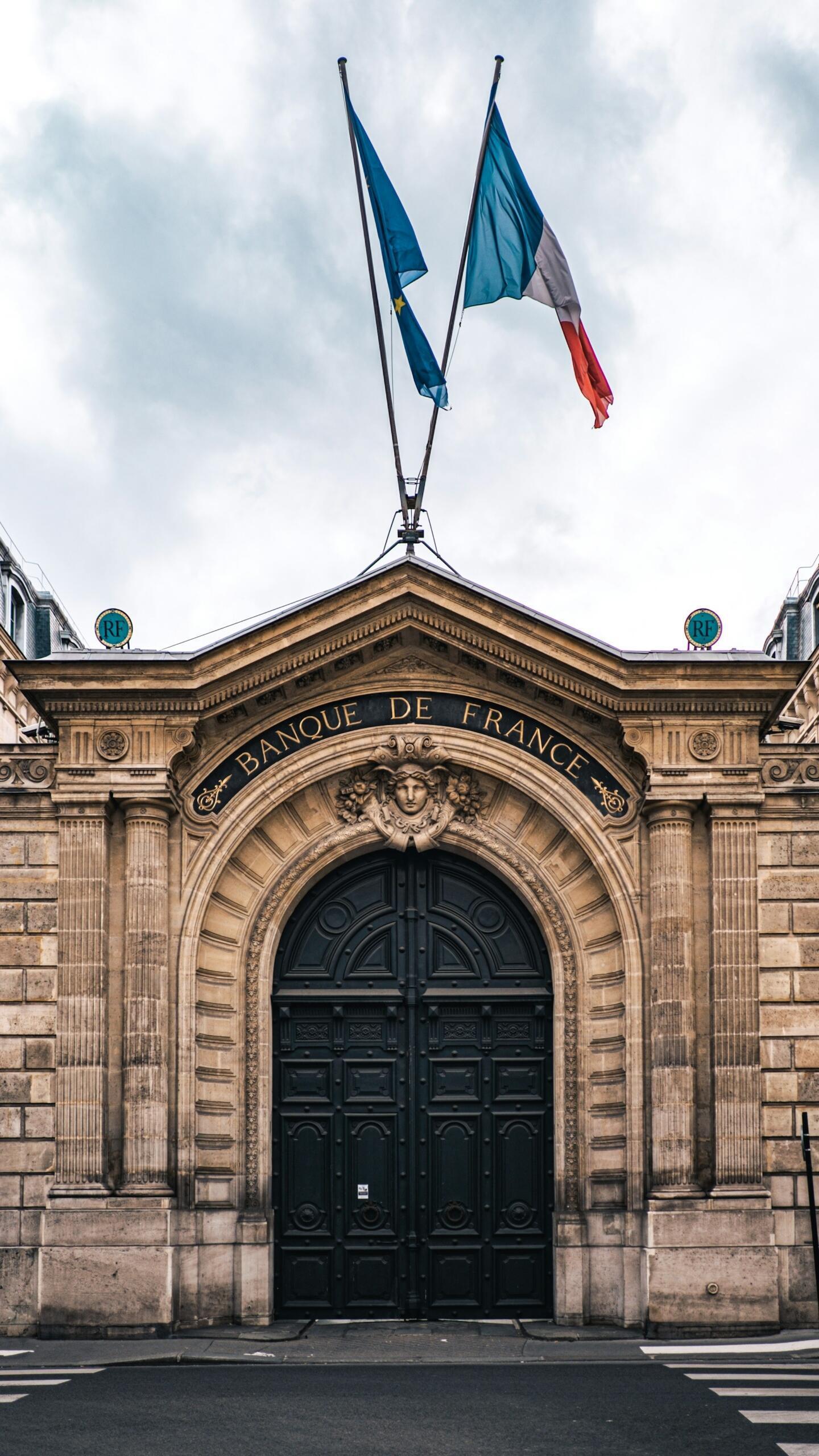 Entrance to the Bank of France, featuring an ornate black gate, classical stone architecture, and the flags of France and the European Union flying above.