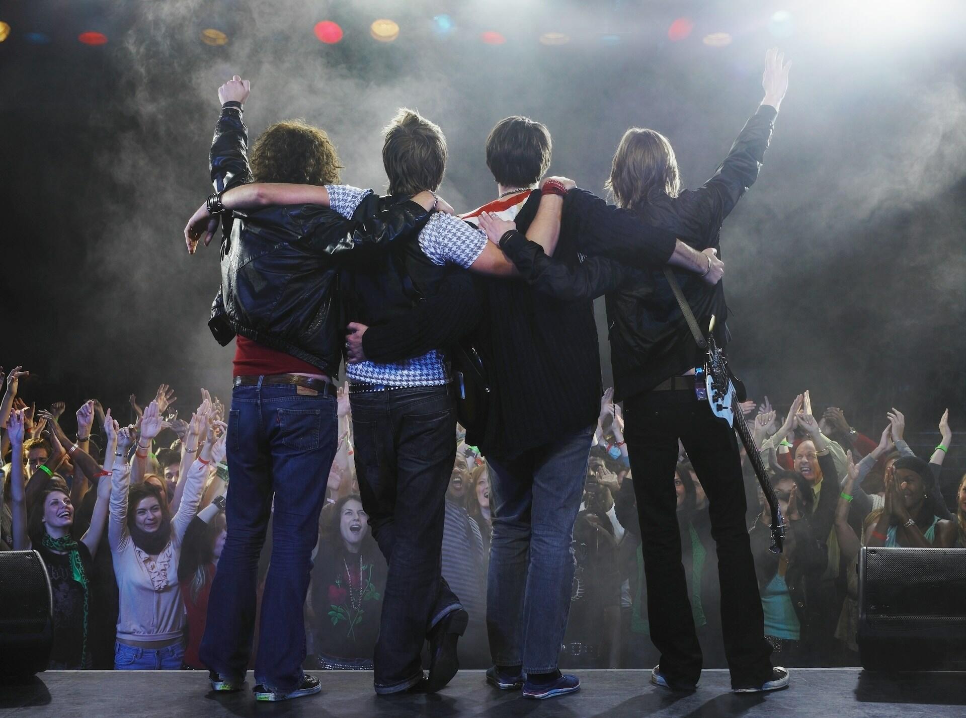Four musicians with the hands in the air facing fans on stage.