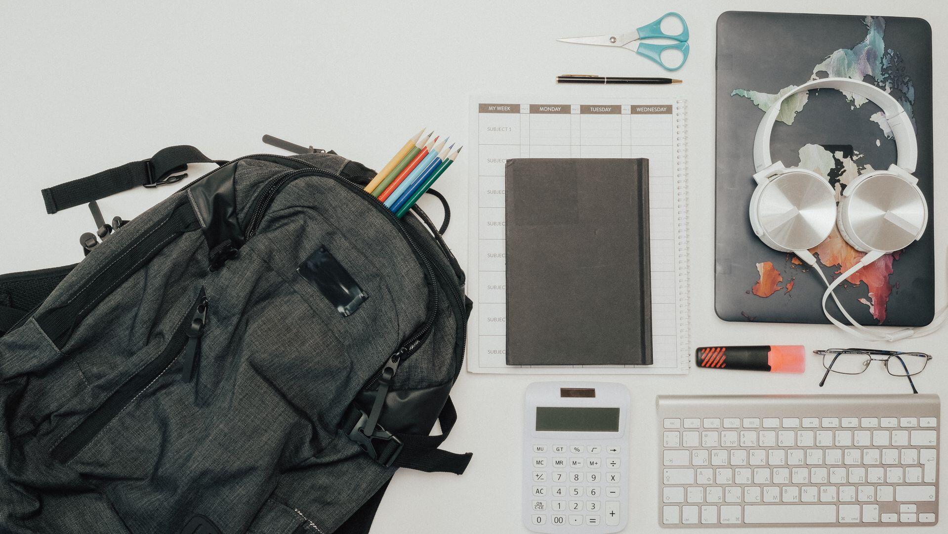 backpack with contents arranged nearby, including a laptop, keyboard, headphones, scissors, highlighter, and colored pencils