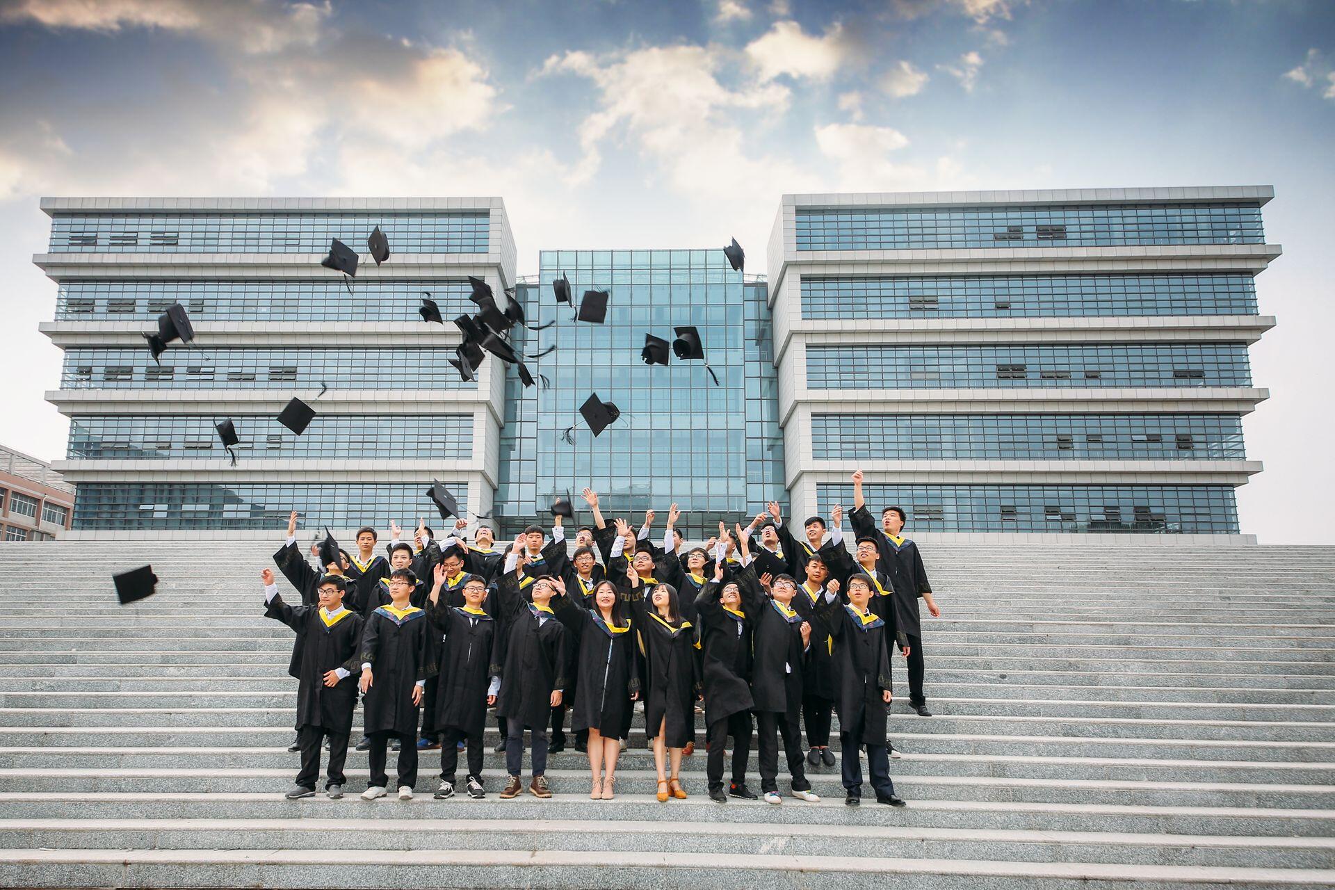 students standing on steps in front of modern college building, wearing gowns and throwing their caps in the air