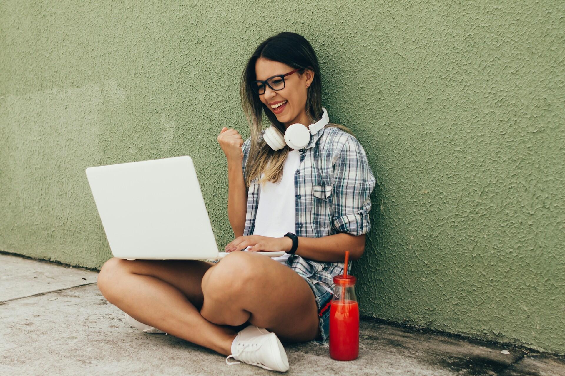 student sitting on the ground and fist pumping while smiling and looking at laptop screen