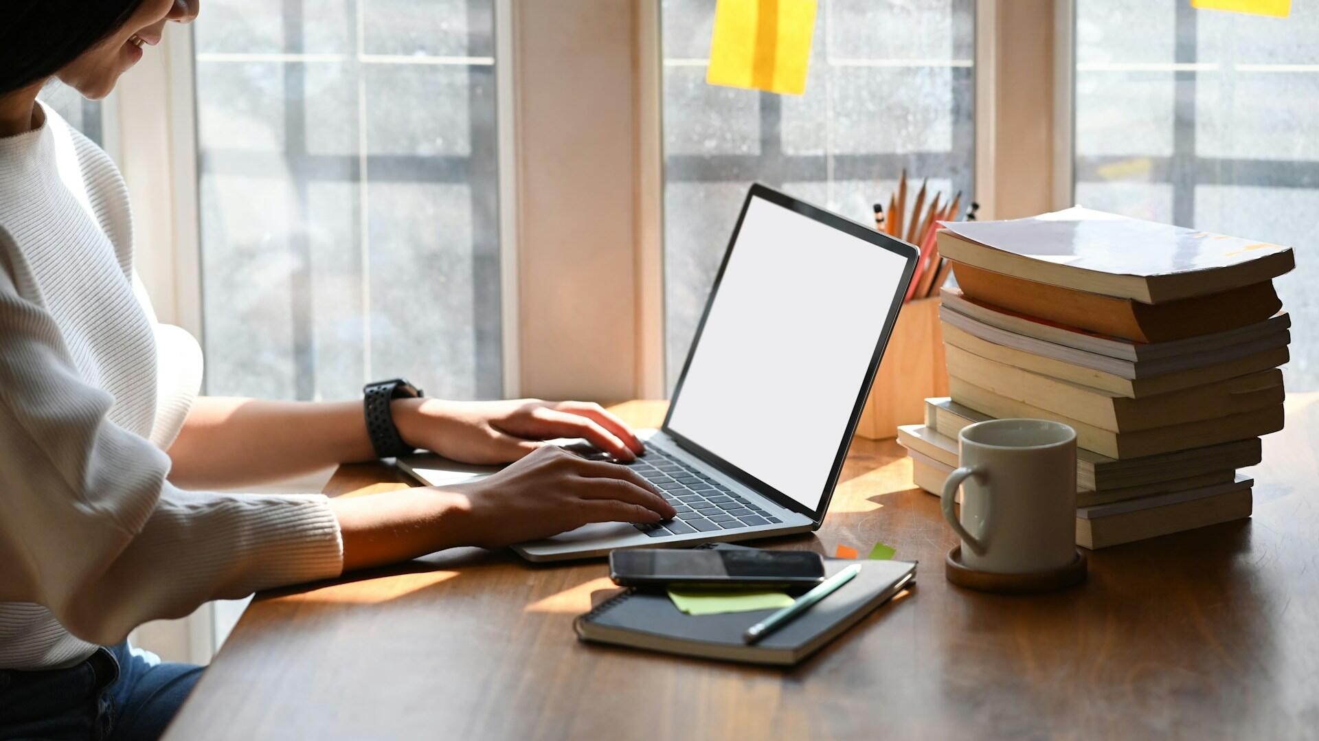 person using a laptop computer on a wooden desk with many books in a well-lit room
