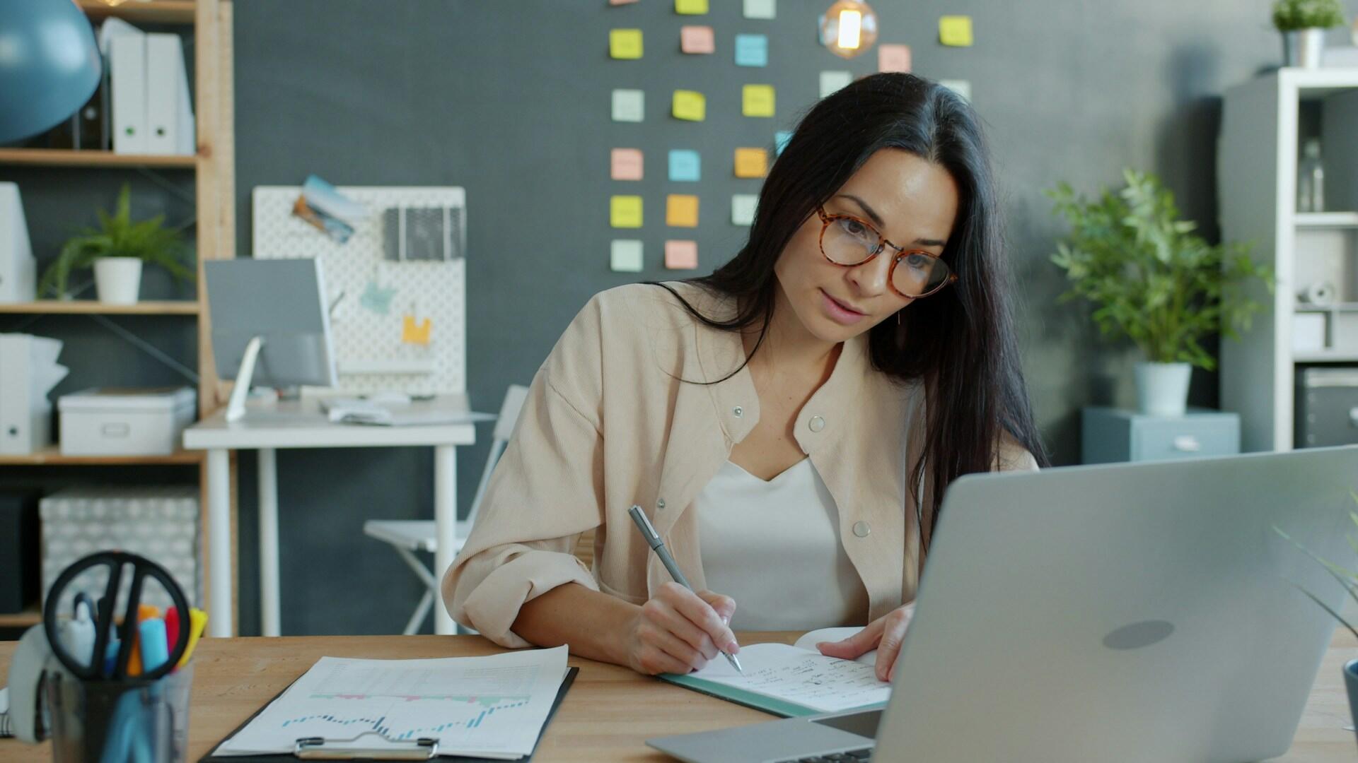person with an organized schedule on their wall writing notes while looking at a computer screen