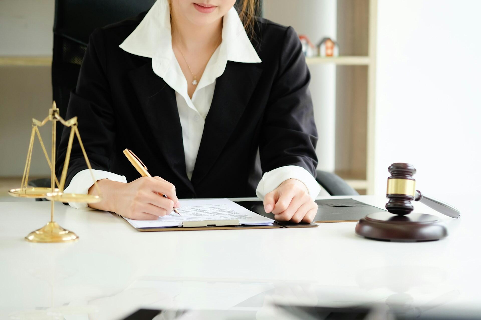Lawyer signing legal documents on their desk. A gavel and decorative scales rest nearby.