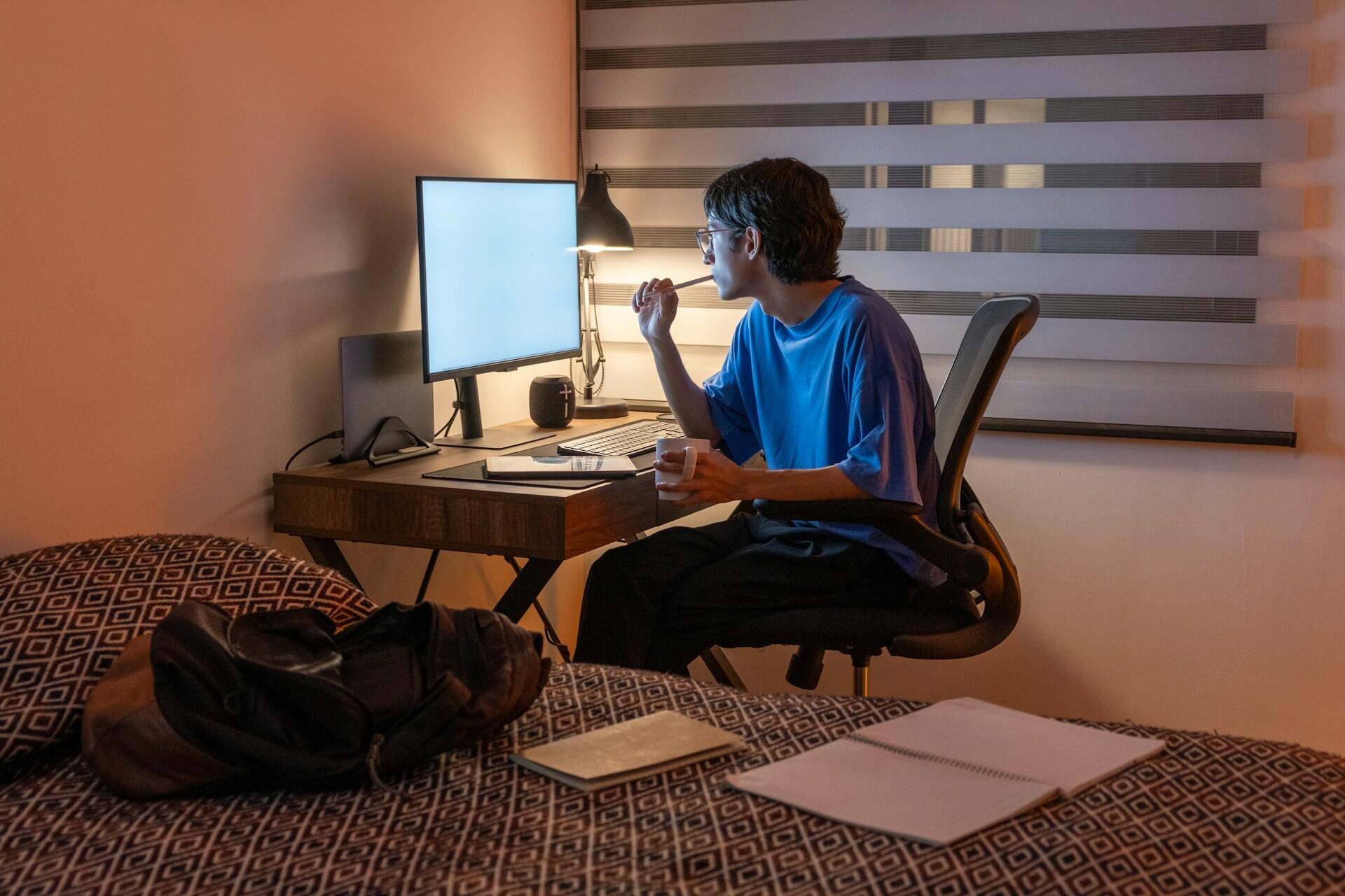 college student studying late at night at a desk with a large computer screen. books and notebook lay open nearby on the bed.