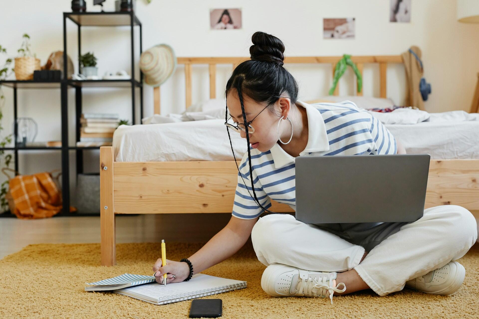 a student sitting on the floor writing notes by hand while looking at a laptop