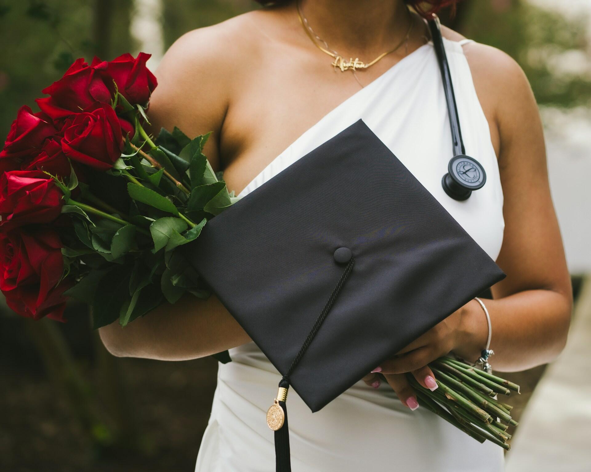 a doctor at their graduation ceremony, wearing a white dress and holding a bouquet of red roses, their graduation cap, and a stethoscope