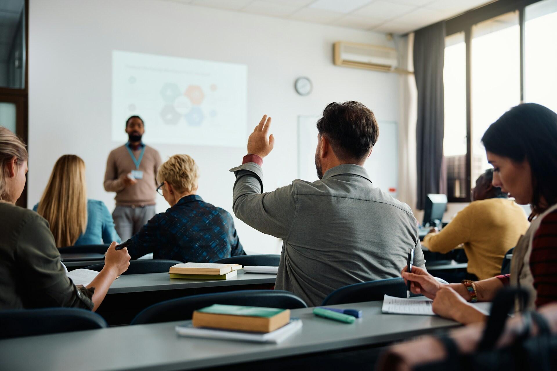 A classroom full of students with one raising their hand, as the professor presents a lesson at the front of the room