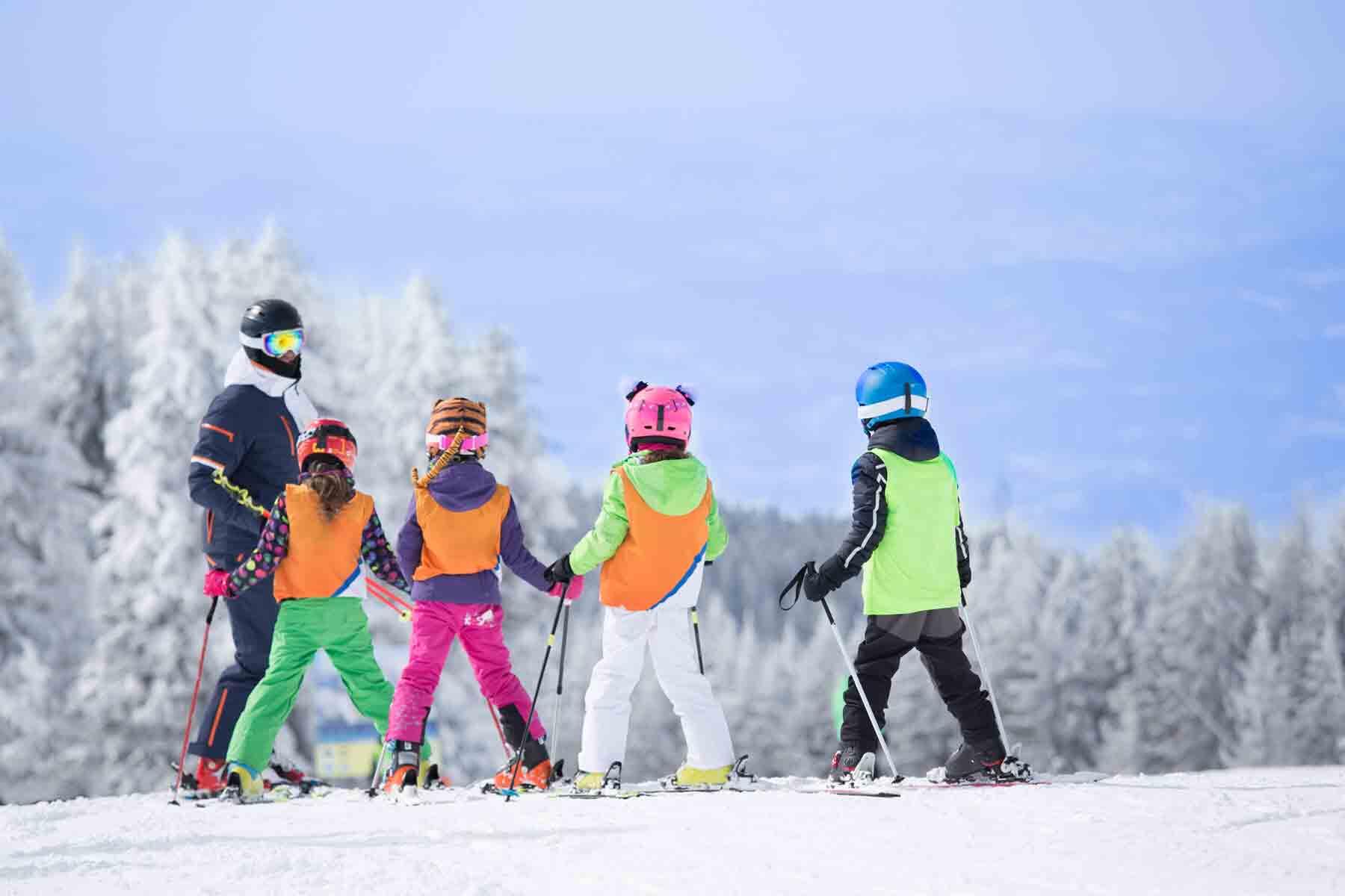 Ski instructor leading four children in helmets and bright bibs across a snowy slope