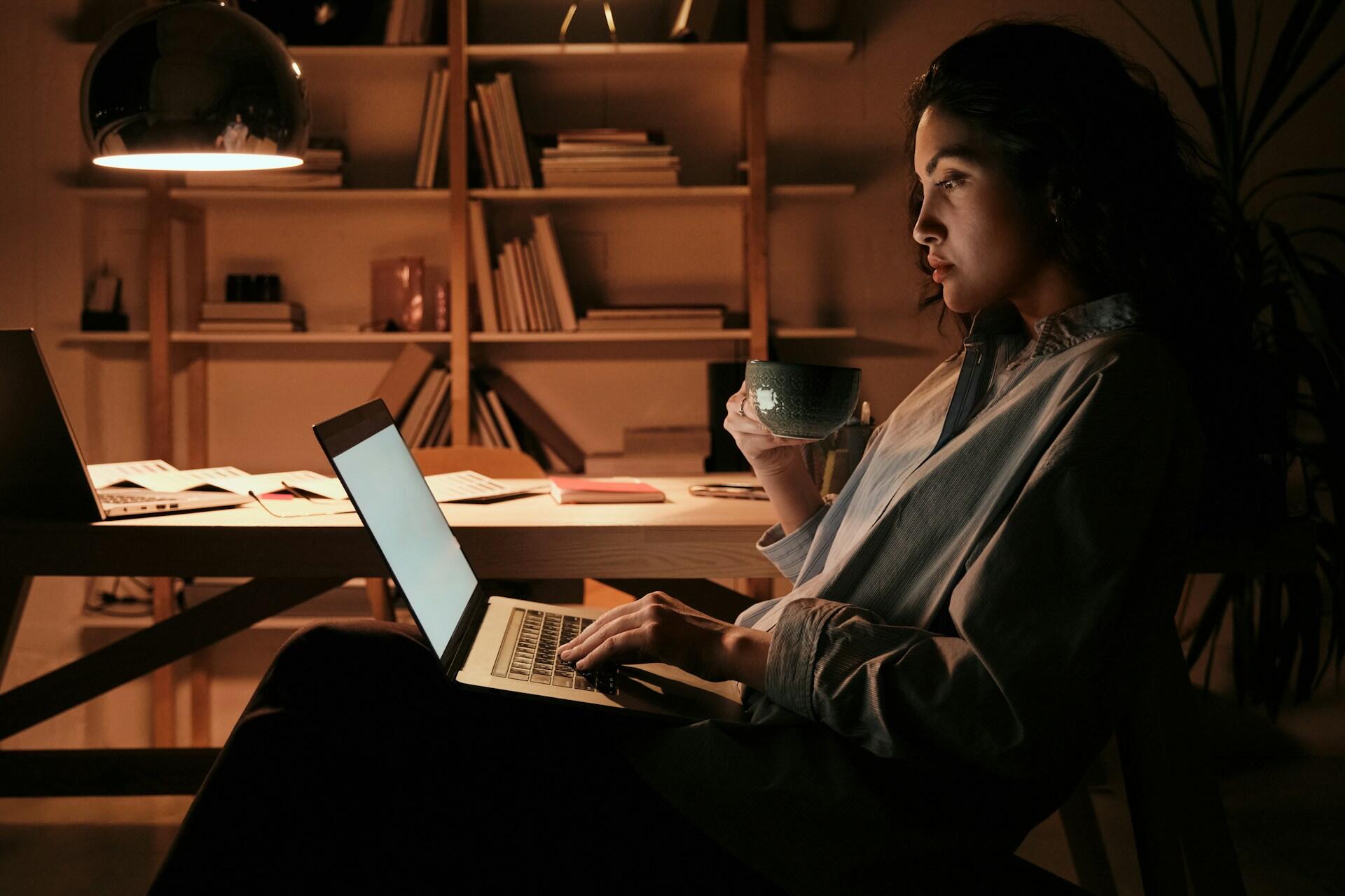 Student reading information on their laptop while holding a cup of tea at night. A desk with additional textbooks is in the background.