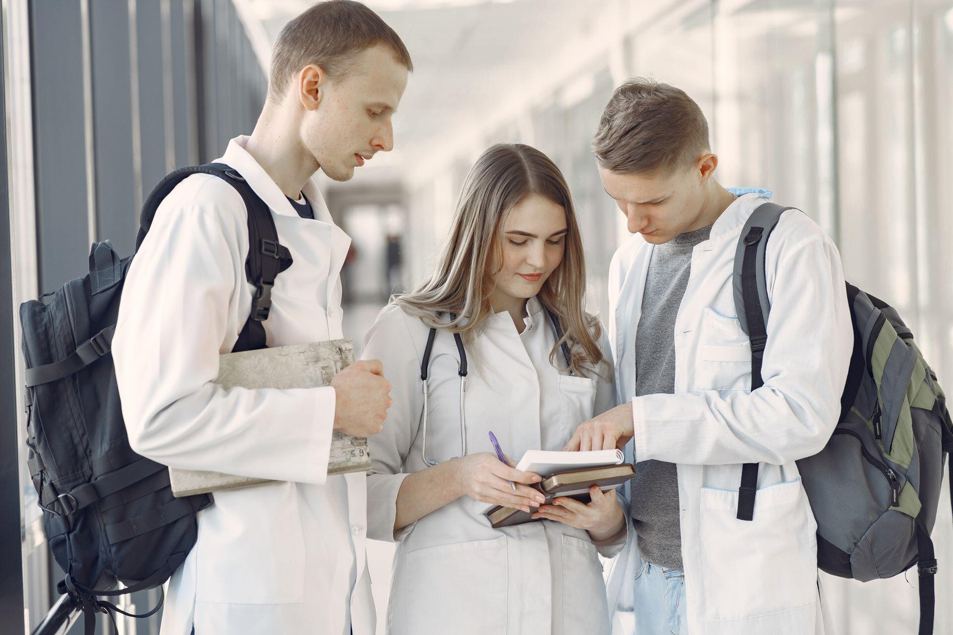 three medical students wearing white coats and stethoscopes sharing notes in a hallway