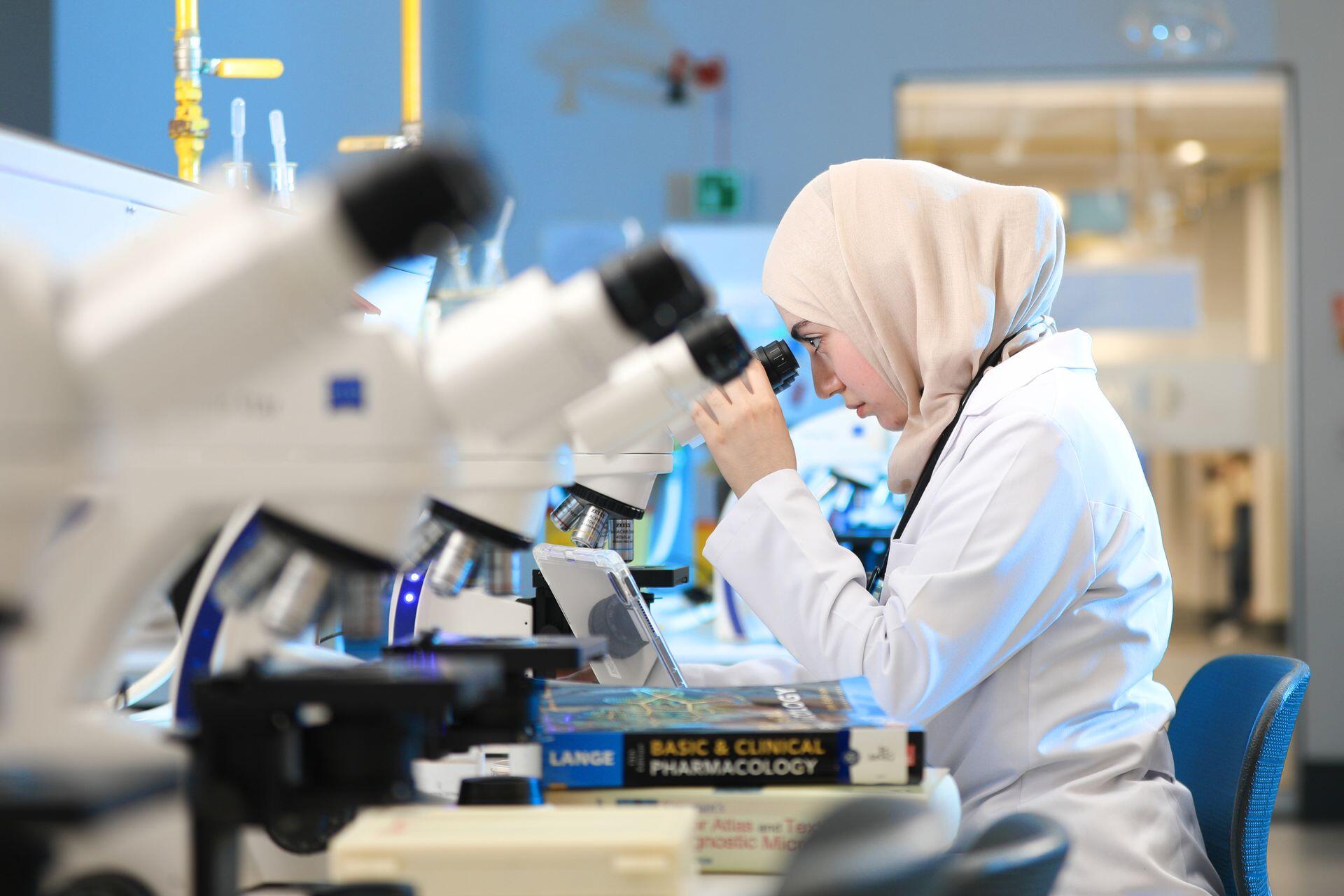 a medical student looks into a microscope in a laboratory classroom with pharmacology textbooks nearby