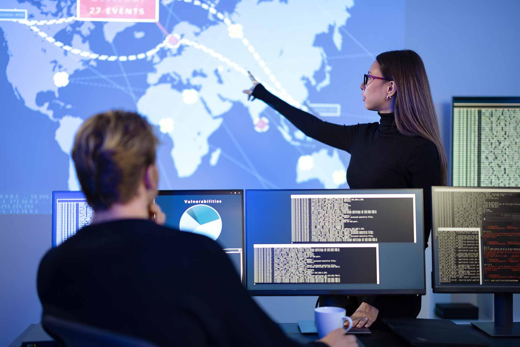 Two cybersecurity analysts reviewing a world threat map and vulnerability dashboards on large screens