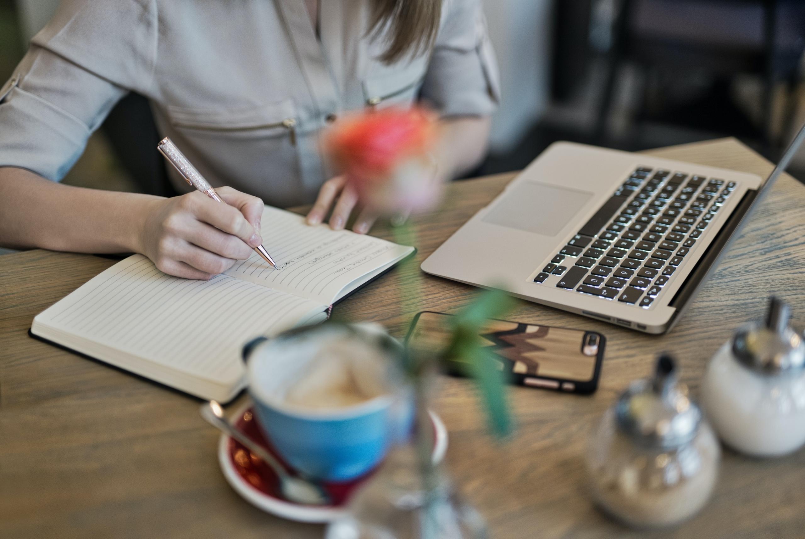 persona escribiendo en un cuaderno con computadora y taza de cafe