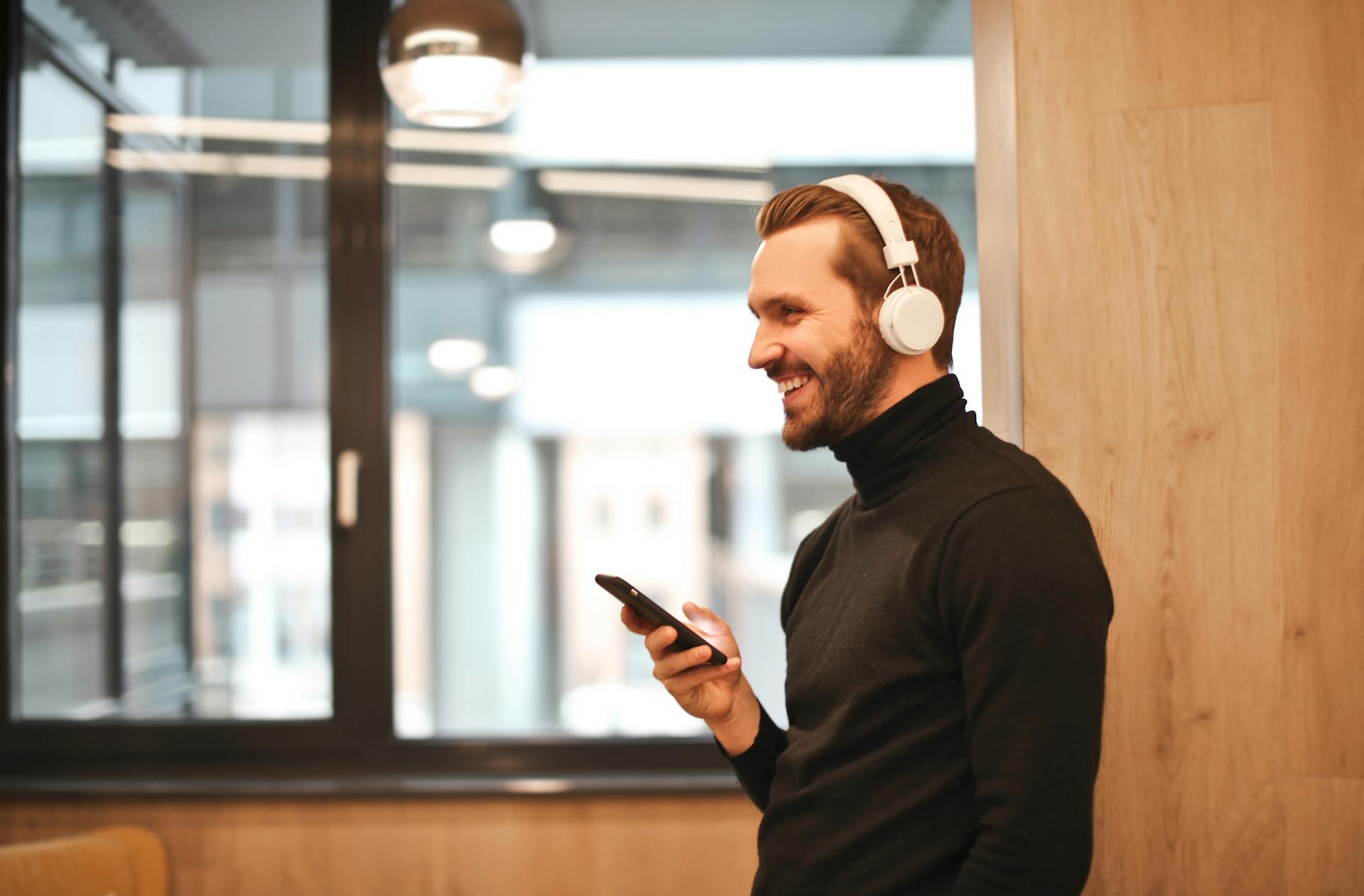 Hombre sonriente con remera negra y auriculares blancos sosteniendo un celular.