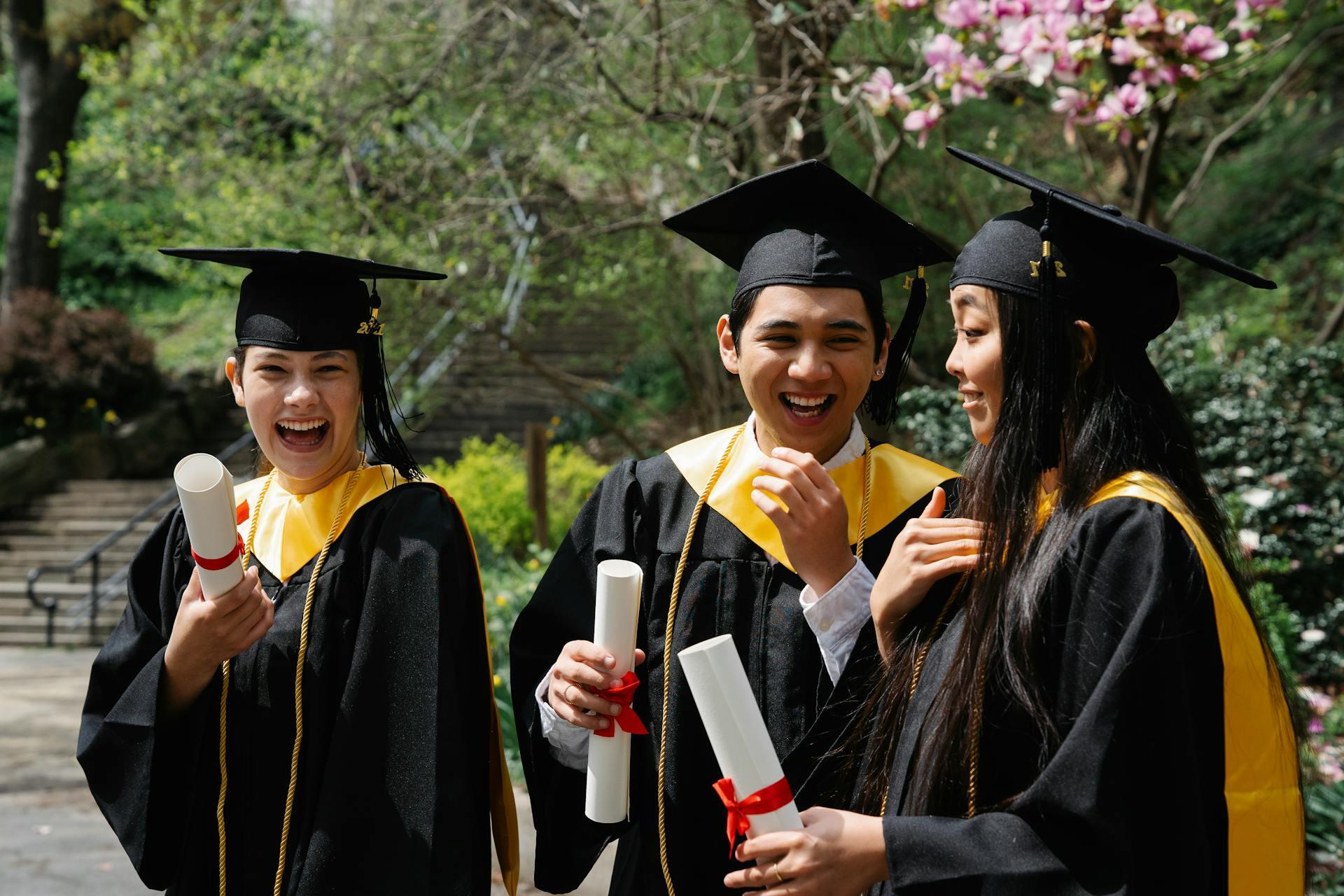dos chicas y un chico vestidos con ropa de graduación festejando con diplomas en la mano