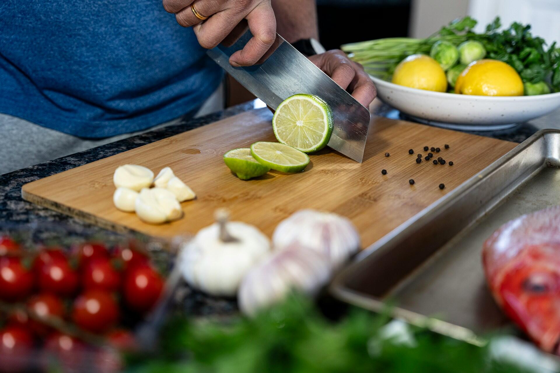 Ua persona prepara una comida cortando una lima sobre una tabla de madera, rodeada de ingredientes frescos como ajo, tomates y pescado. Foto de Anju Ravindranath en Unsplash.