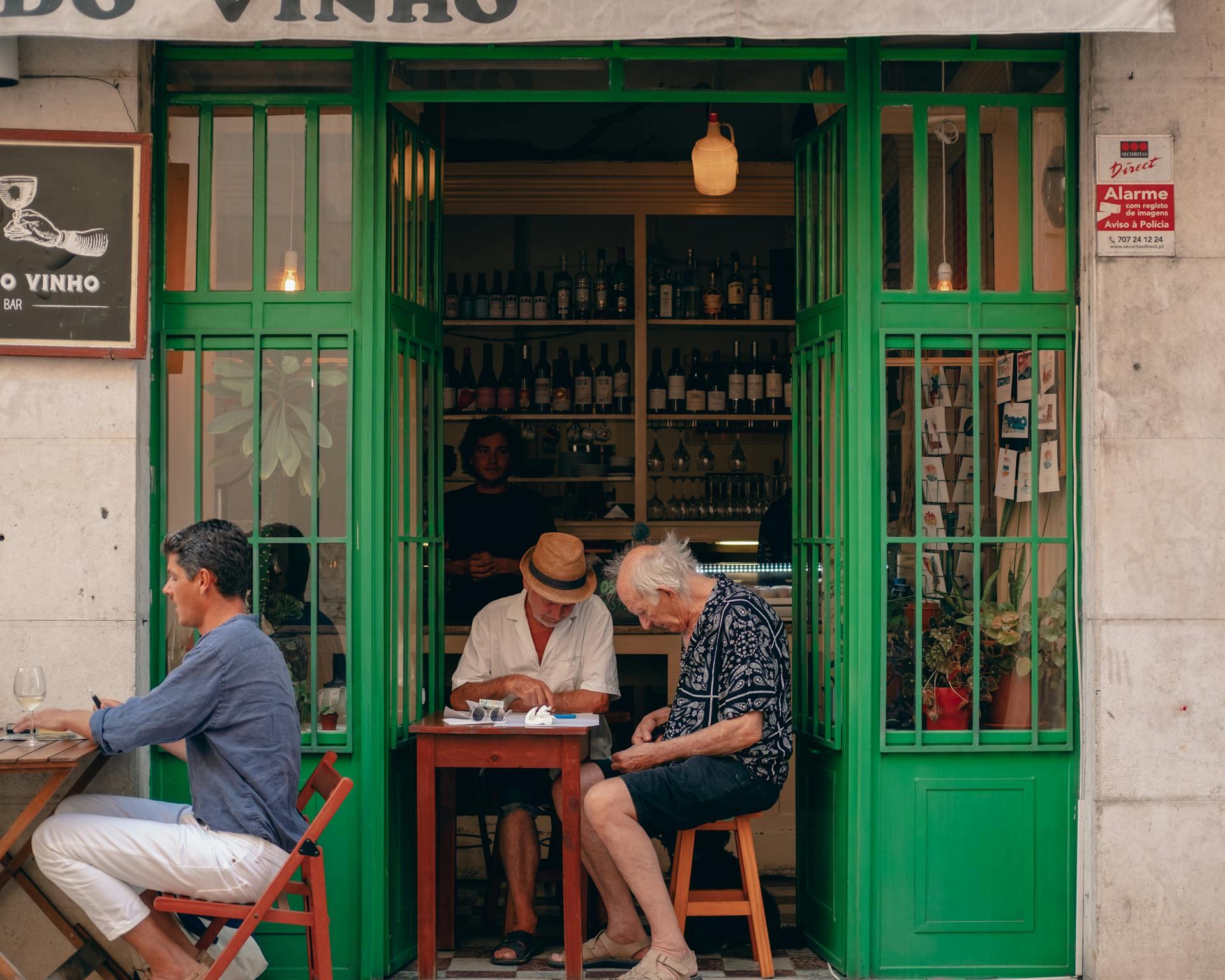 Tres hombres disfrutan de una bebida en la vereda de un bar de vinos con fachada verde en Lisboa, foto de Mucahit Tutuncu en Pexels.