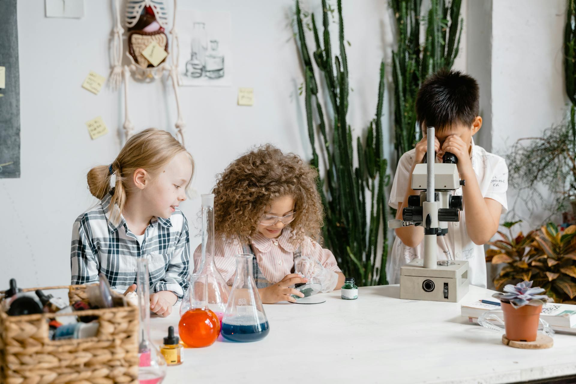En un aula de ciencias, tres niños pequeños colaboran en una mesa; uno mira por un microscopio mientras los otros dos observan frascos de laboratorio. Fotografía de MART PRODUCTION.